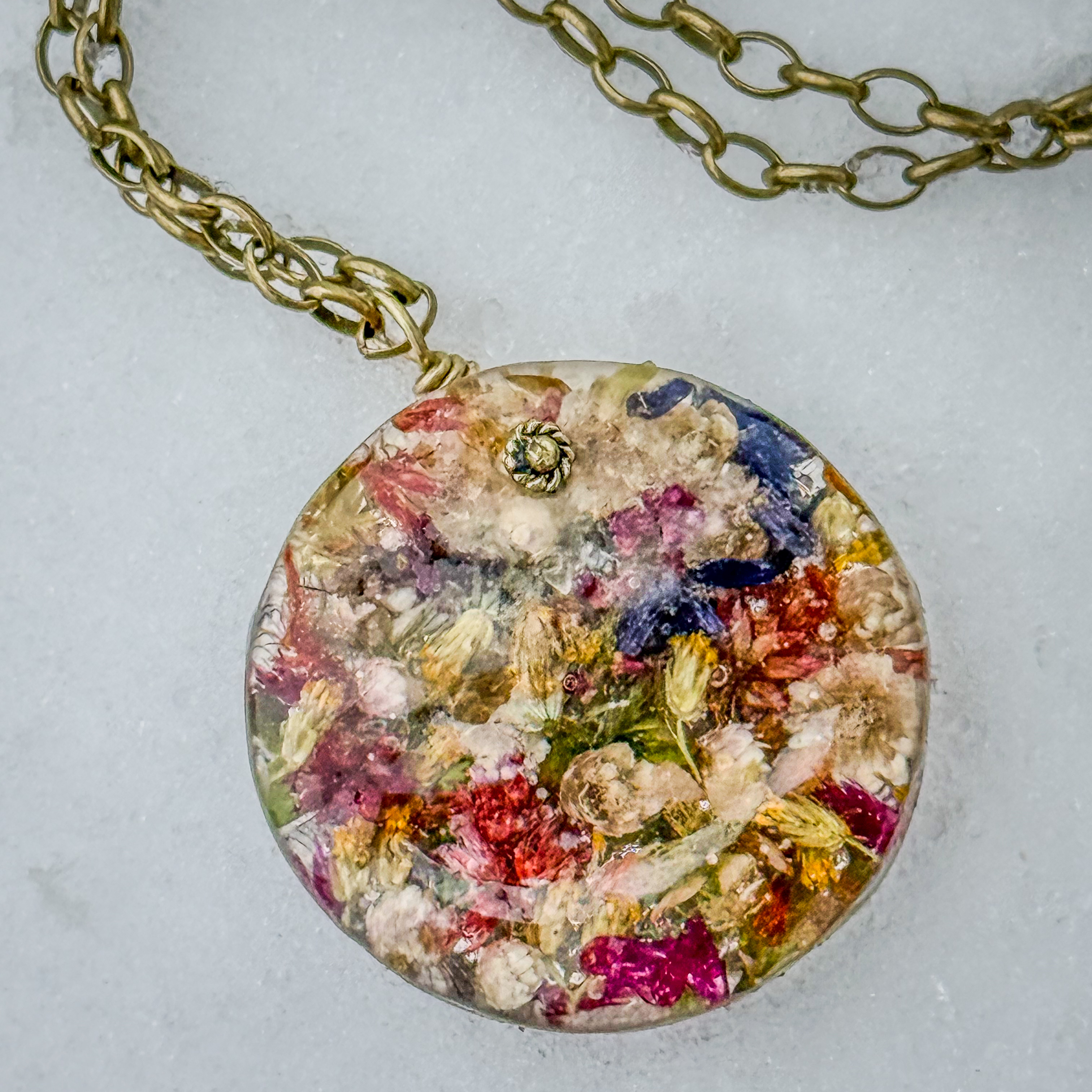 Large round resin pendant on a gold chain, densely filled with pixelated botanical material in dozens of colors — like a miniature Impressionist garden
