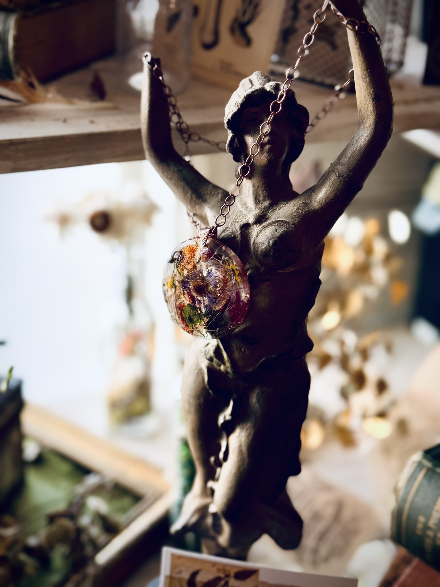 A bronze figure holding a botanical pendant upward, toward the light — the human form and the made thing, together