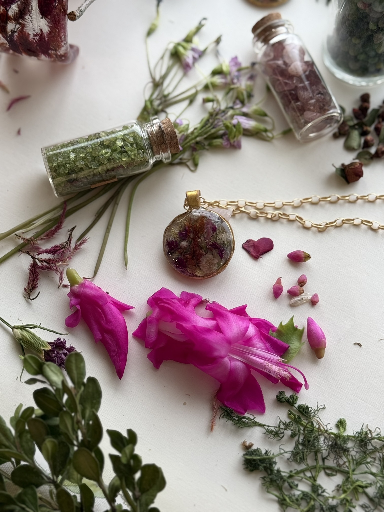 A round botanical pendant on gold chain beside vivid magenta Christmas cactus blooms and crystal-filled vials