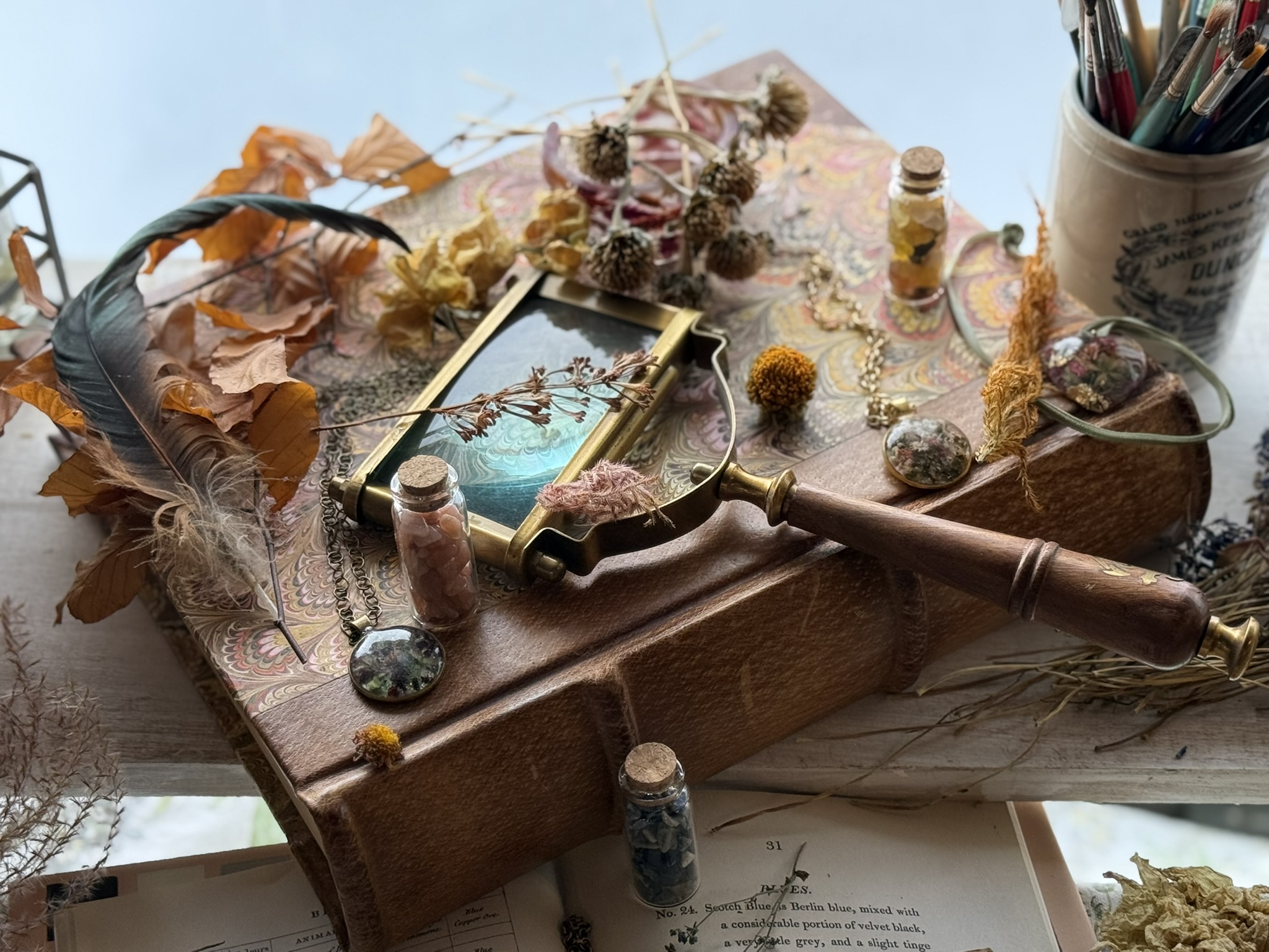 Pendants and vials on a leather-bound reference book with magnifying glass — the studio at work