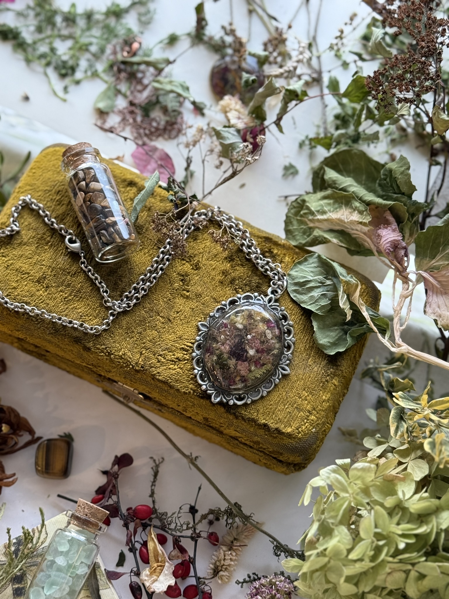 A silver ornate pendant resting on an ochre velvet box, surrounded by hydrangea and seed-filled vials
