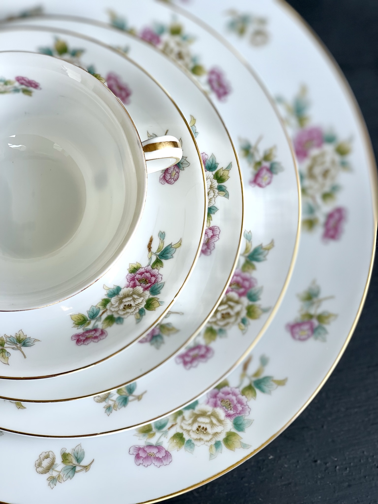 A cup and saucer with pink peonies and cream roses on white, olive-teal leaves, gold rim — the title piece of the collection, shown nested on a plate and saucer