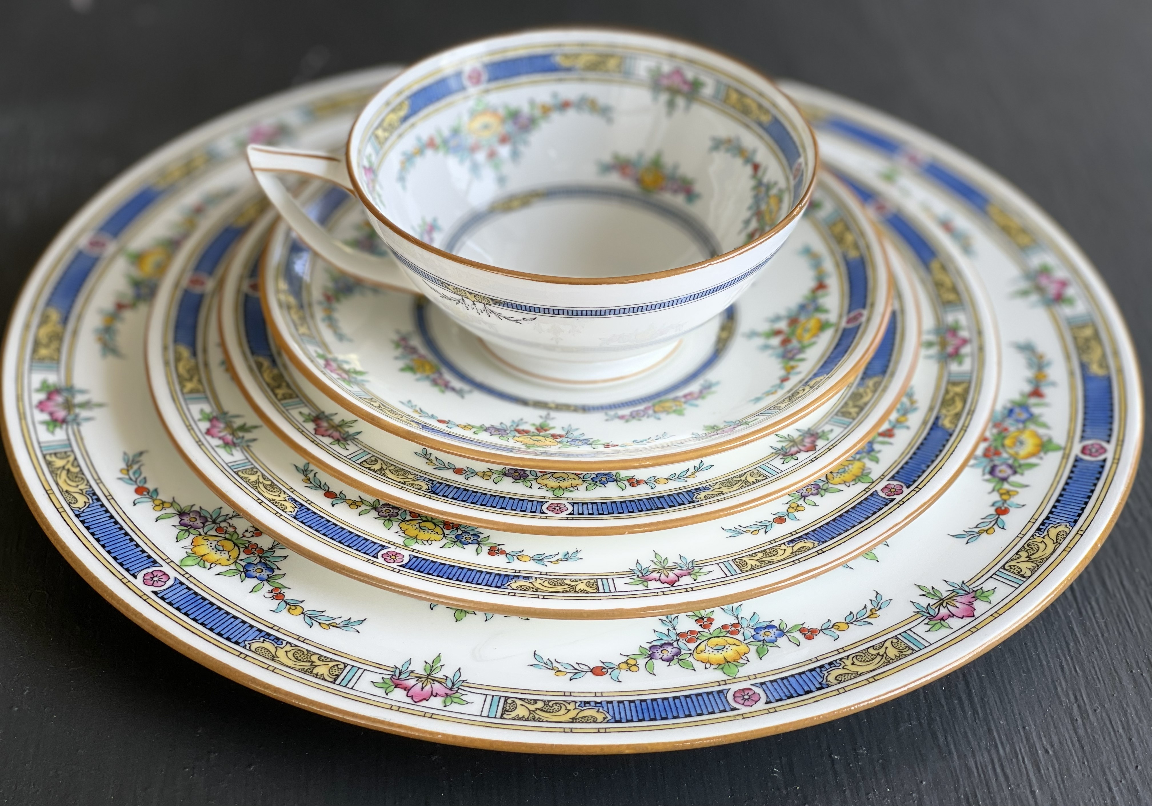A complete five-piece place setting fanned out concentrically — dinner plate, salad plate, bread plate, saucer, and teacup all nested together in cobalt blue and gold