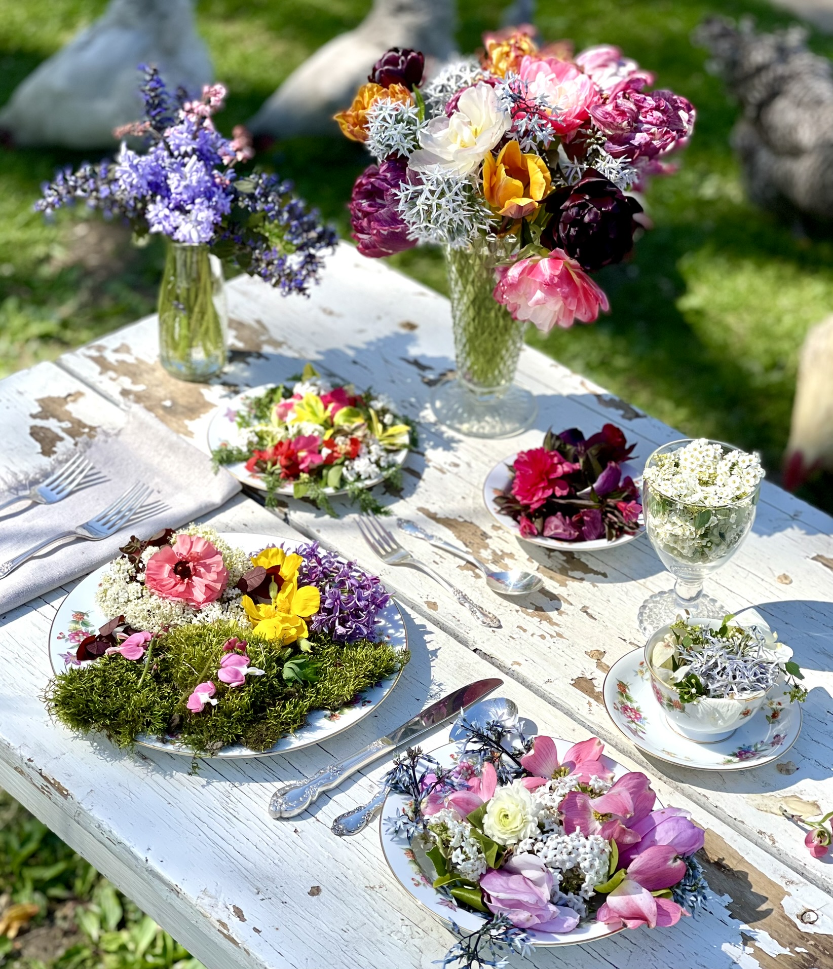 Overhead angle of vintage china plates filled with living garden flowers on a weathered white outdoor table — the meeting of two obsessions