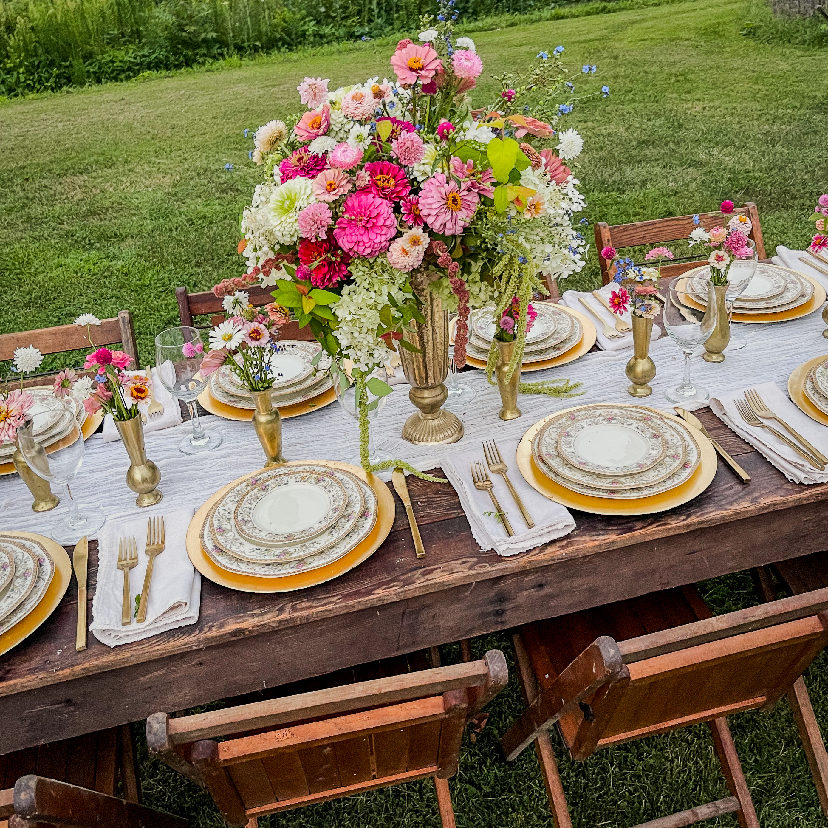 Wide overhead of the formal farmyard feast table — gold chargers, brass bud vases, a towering centerpiece of garden flowers