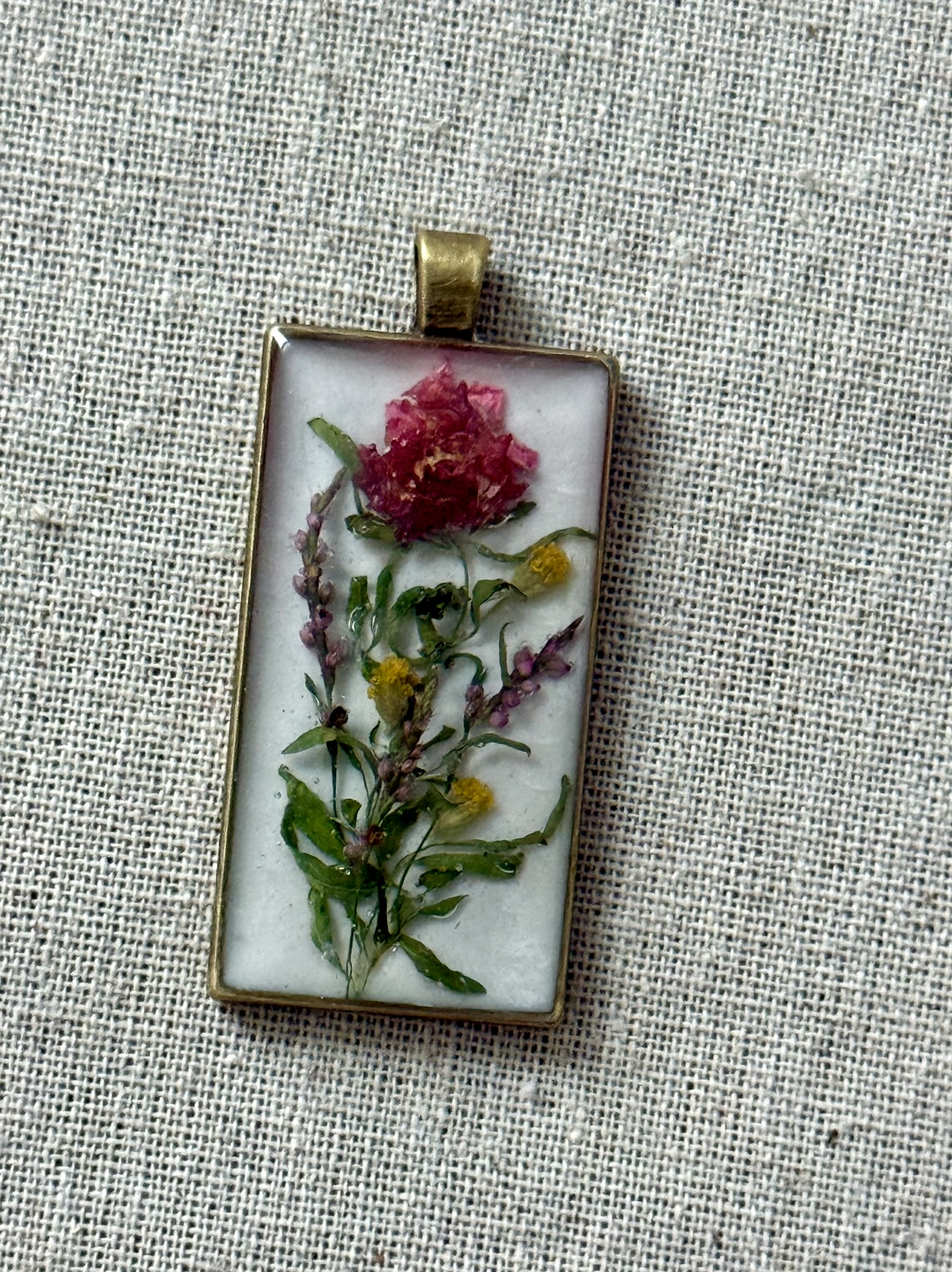 Rectangular brass pendant: a crimson lily petal twirled into a rose, standing above a meadow of stems in resin