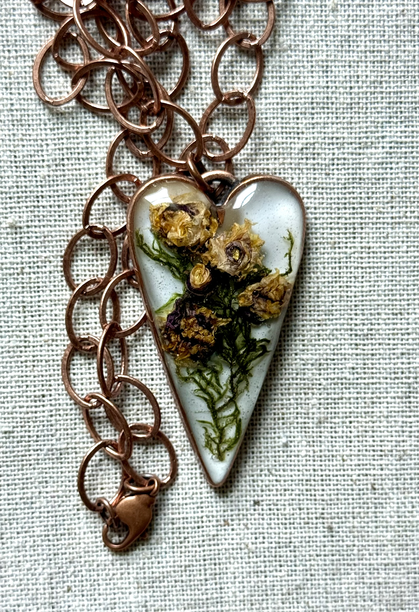 Copper heart pendant on copper oval chain, golden lily-roses and green moss inside, on linen