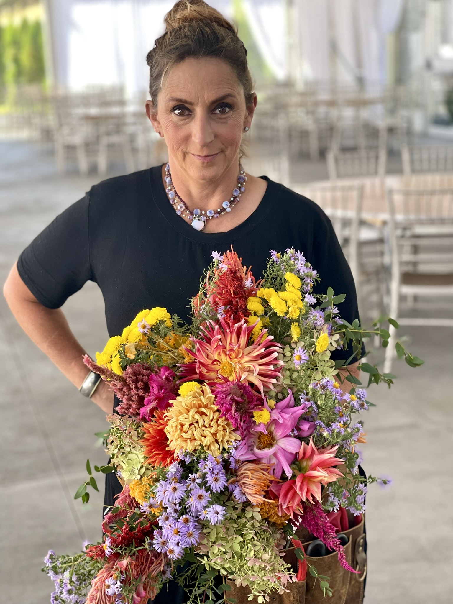Luli in black, floral necklace and leather tool pouch, holding a massive colorful bouquet — dahlias, celosias, asters, marigolds — in a wedding venue tent. The chicken wire bouquet base is hidden inside.