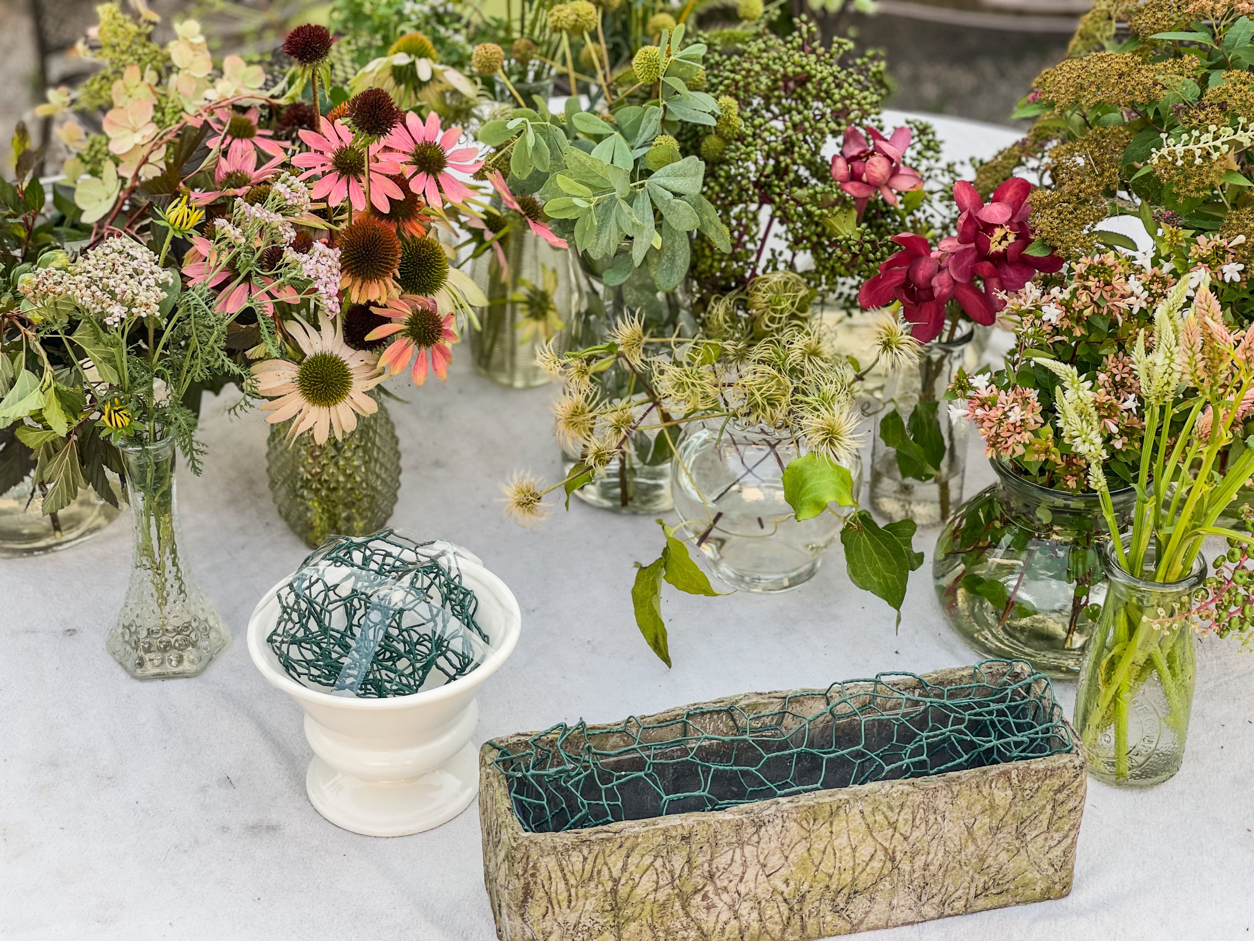 White table with multiple vessels — wire dome frog in white compote bowl, chicken wire in concrete planter, echinacea, clematis, hydrangea, dahlias arranged all around