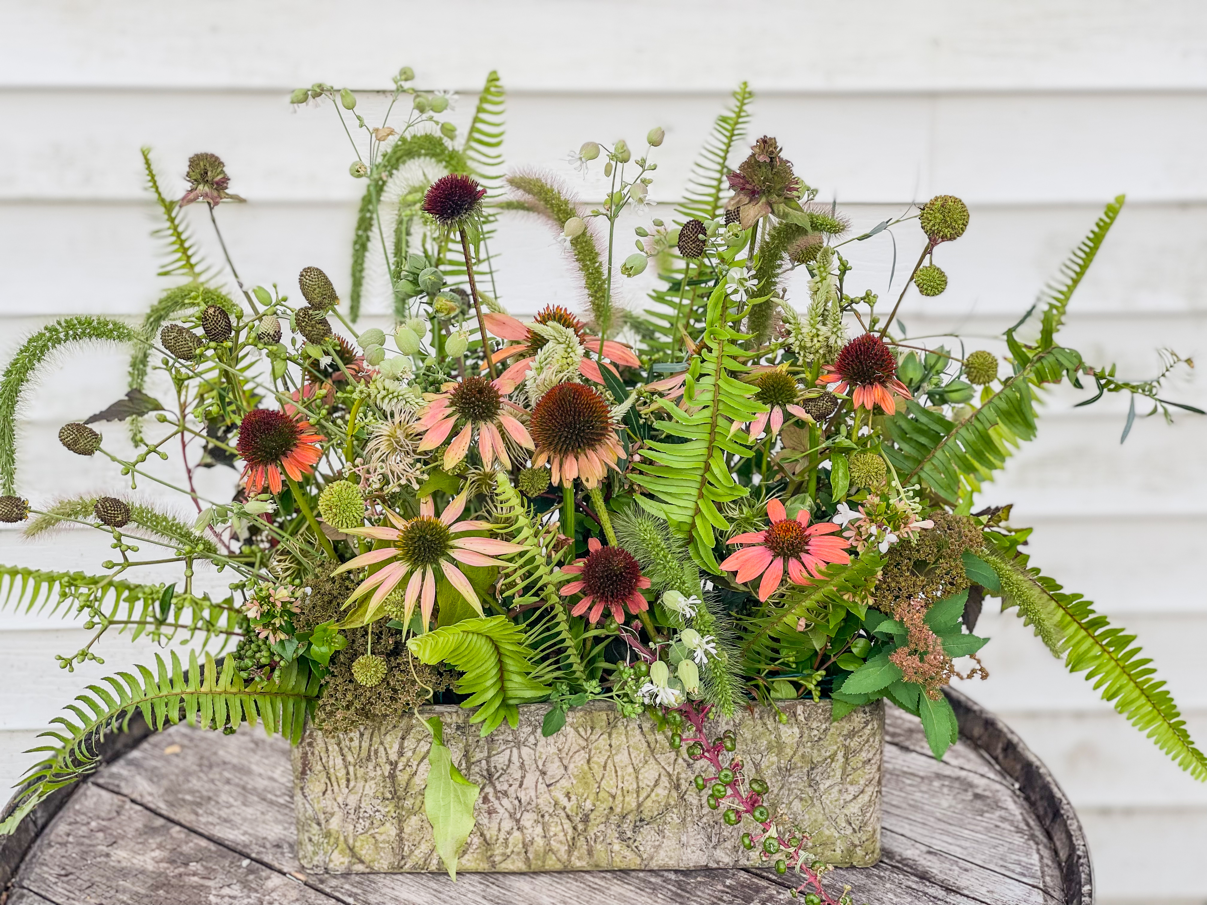 Concrete planter on weathered barrel top against white clapboard — dense foraged arrangement of echinacea, ferns, teasel, globe thistle, veronicastrum, and wild seedheads