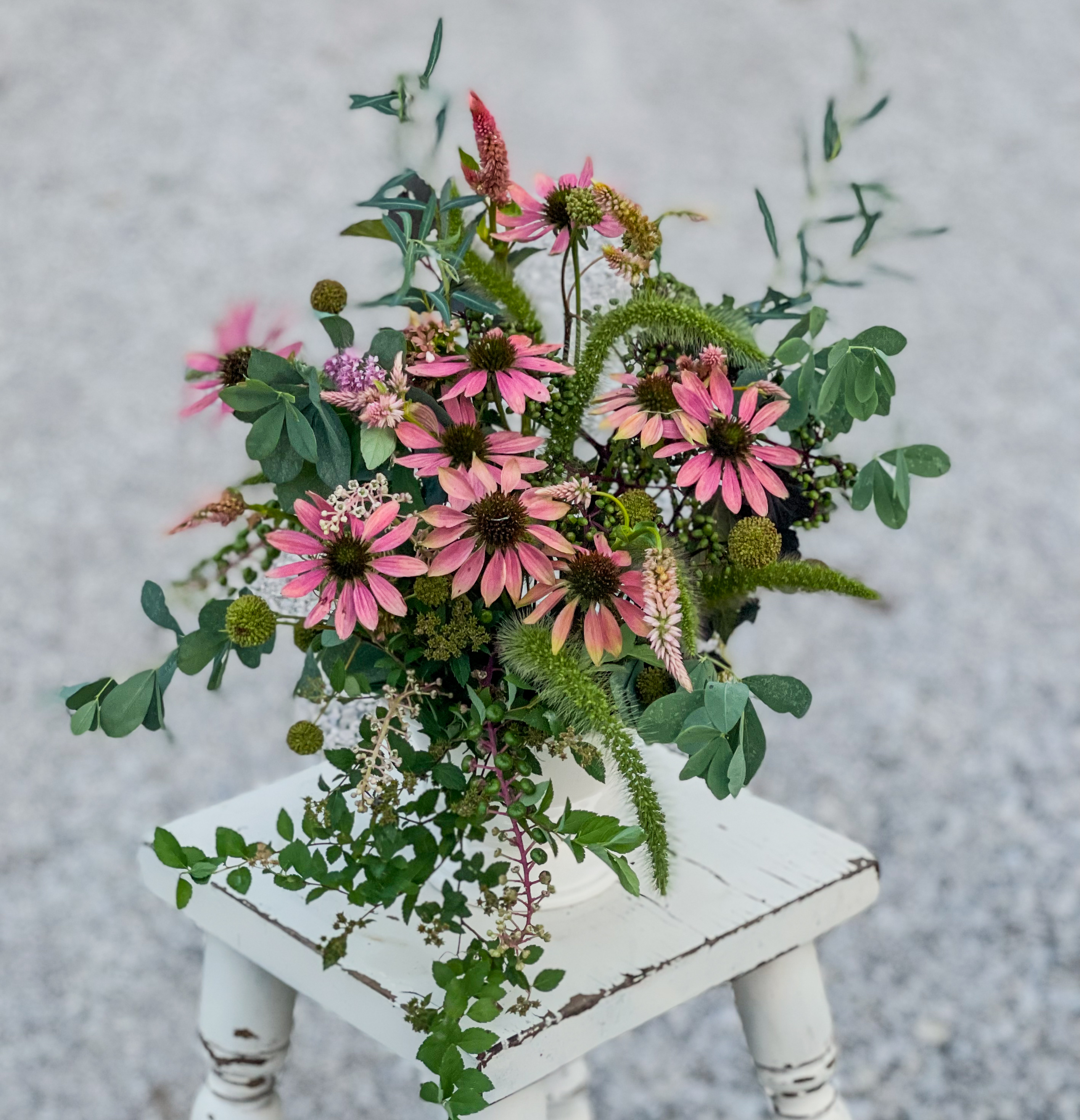 Pink coneflowers, green berry clusters, teasel heads and eucalyptus in a white vessel on a white chippy-paint stool against gravel — the wire frog invisible inside