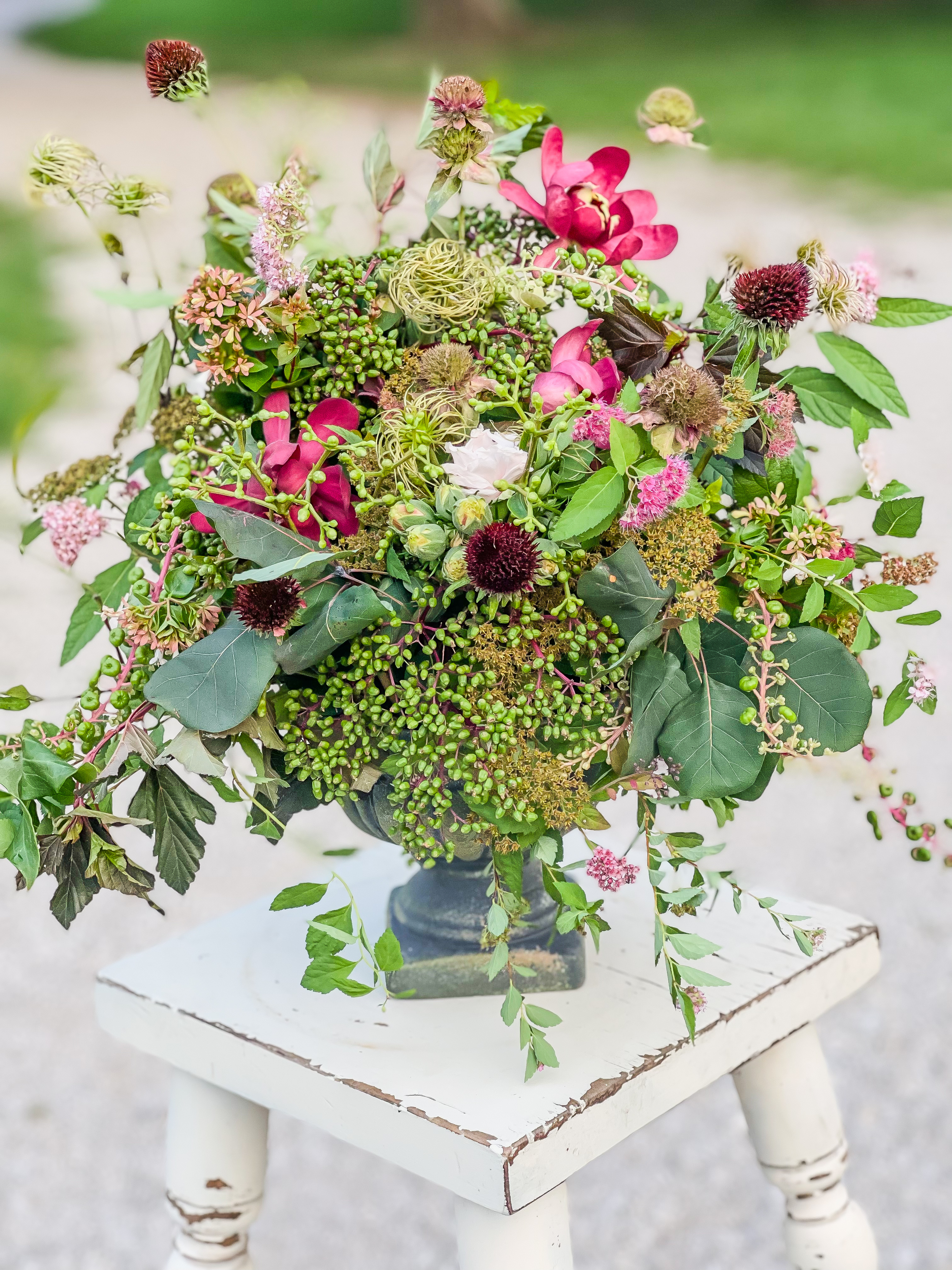 Dark cast iron urn on a white stool overflowing with magenta clematis, green sedum berries, pink echinacea, globe thistle, and trailing foliage — lush and wild