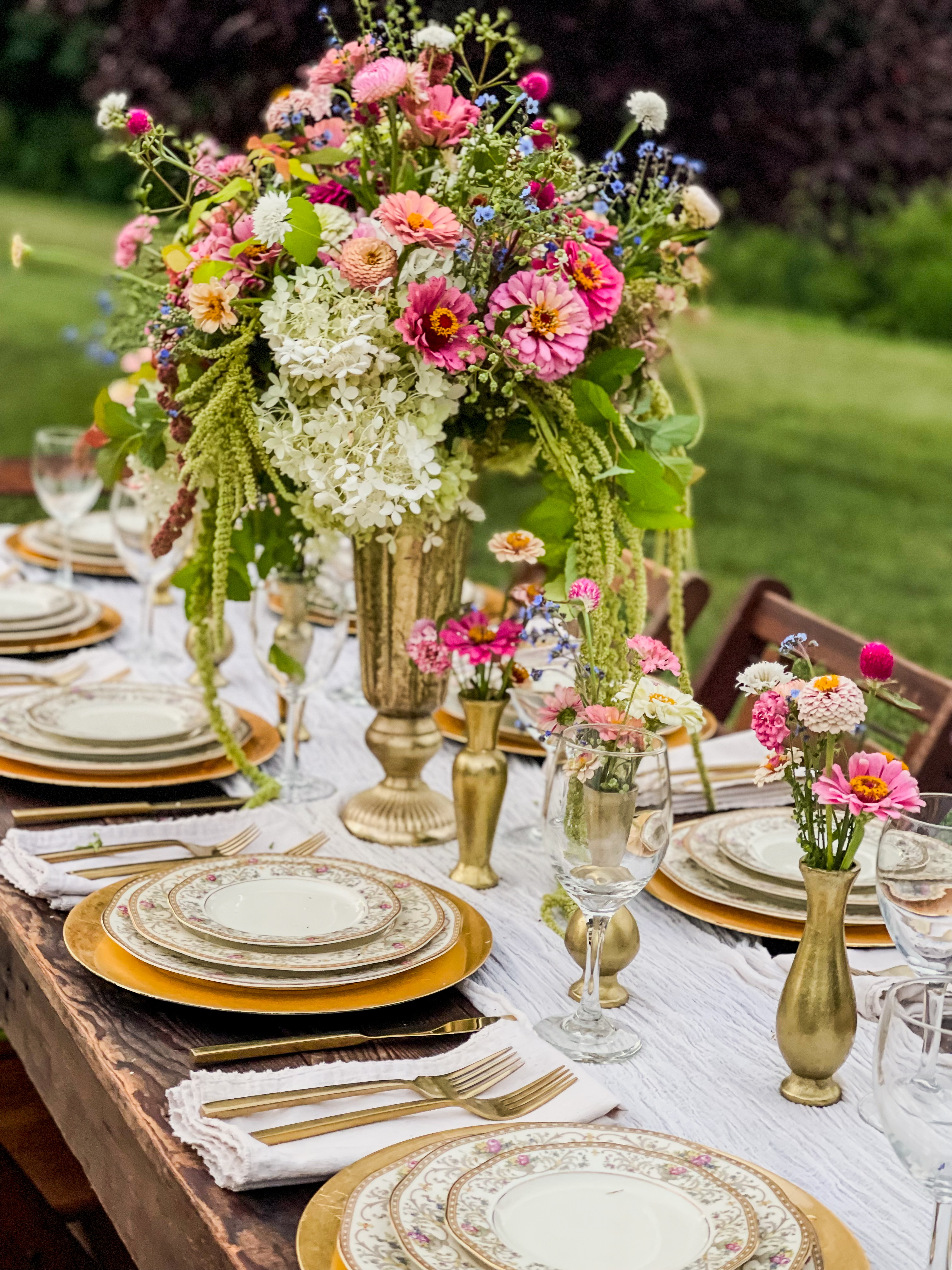 Long outdoor dining table set for a formal feast — brass compote urn overflowing with zinnias, dahlias, hydrangea and amaranth, mismatched vintage china, brass bud vases