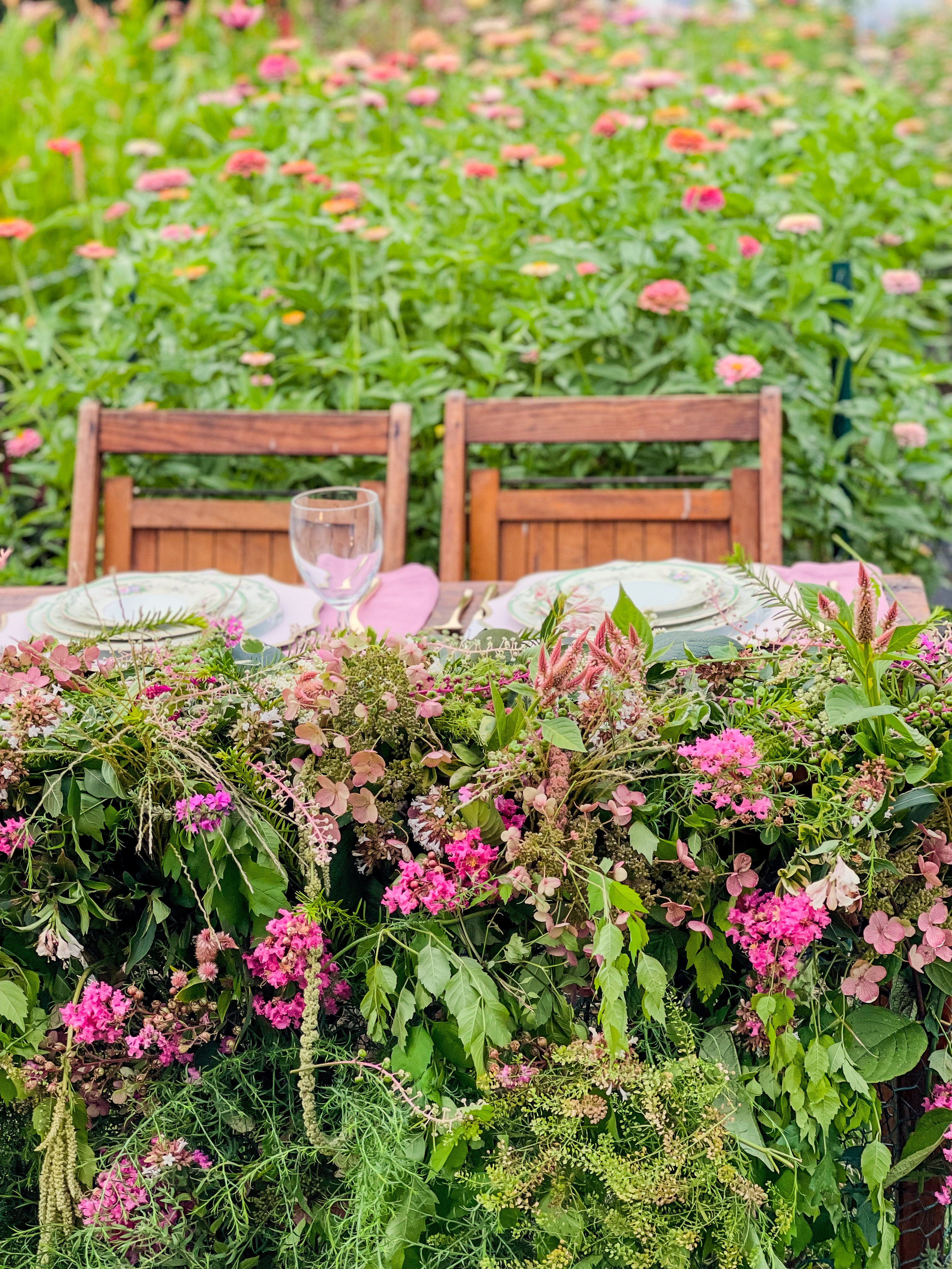 A farmhouse dining table set in the middle of a zinnia field in full bloom — a lush pink and green floral garland cascades over the front edge, two chairs wait, vintage china place settings visible through the flowers