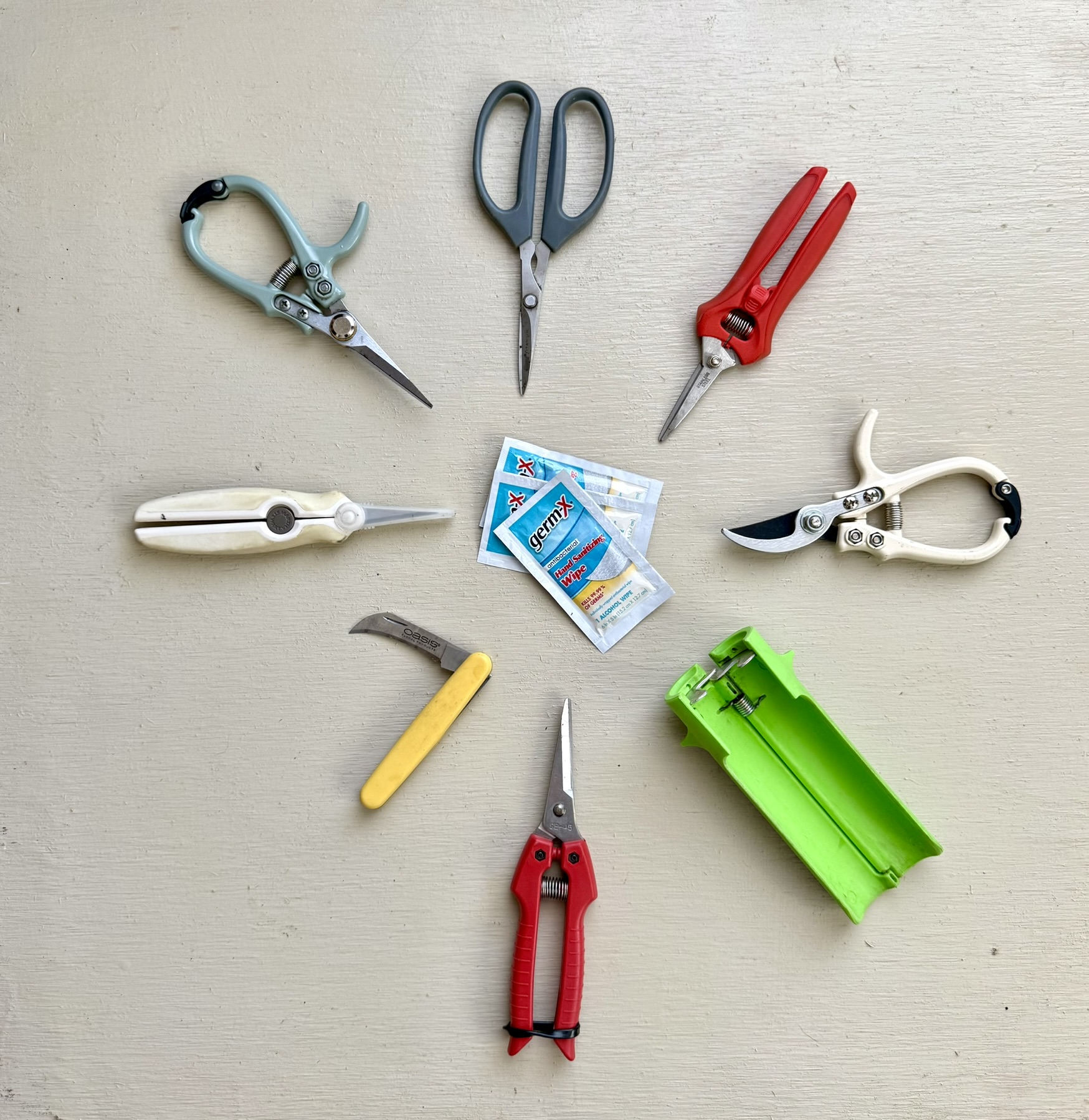 Seven cutting tools arranged in a radial star: floral scissors, bypass pruners, two snips, Japanese precision scissors, OASIS knife, foam cutter — Germ-X wipes at center