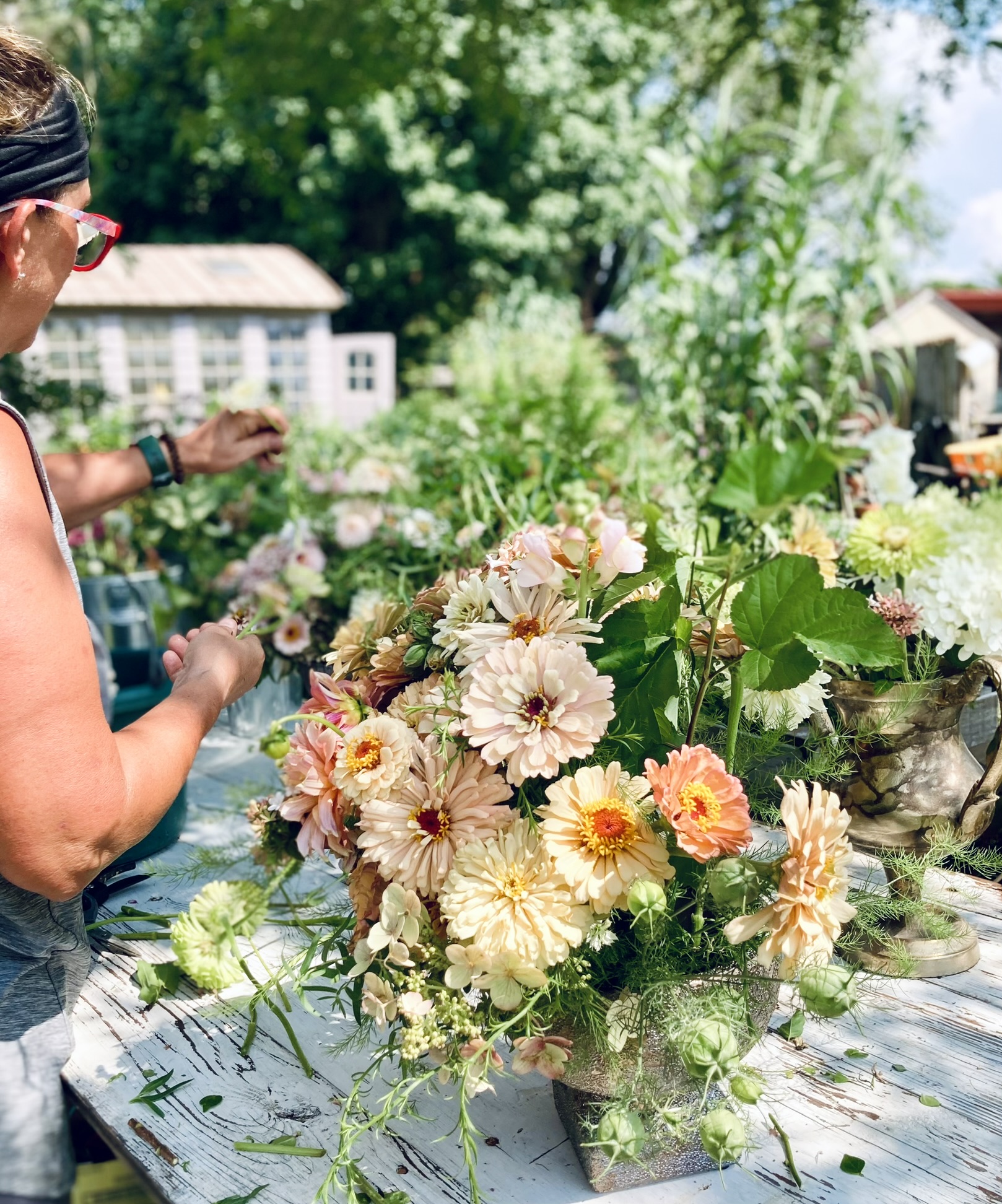 Luli at the outdoor design table building a peach and cream zinnia arrangement in a stone urn — greenhouse visible behind
