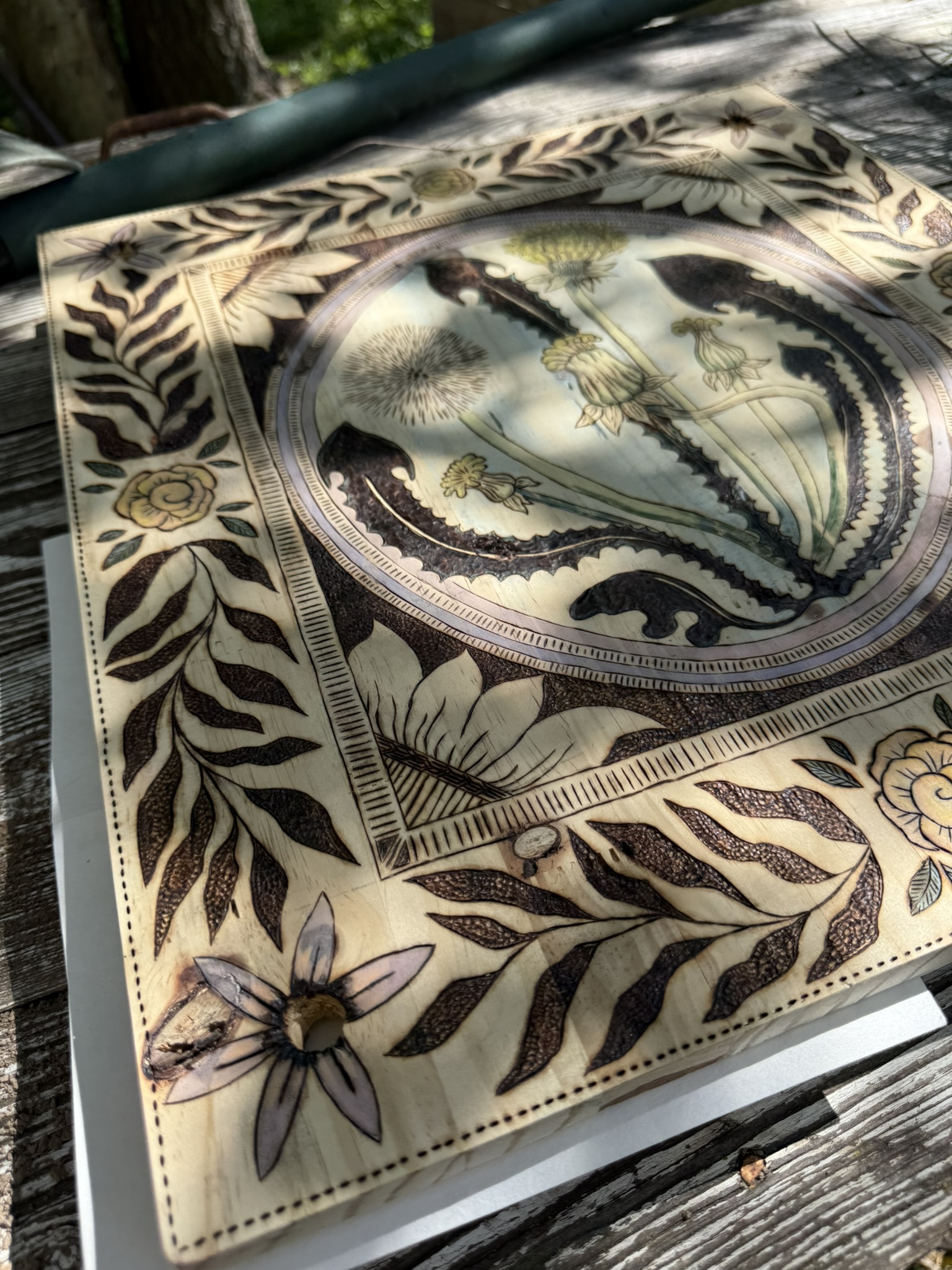 Corner detail of Paige's press — close pyrography work showing the lavender six-petal star bolt detail, vines with hand-colored green leaves, the feather motif, and the triple-ruled border