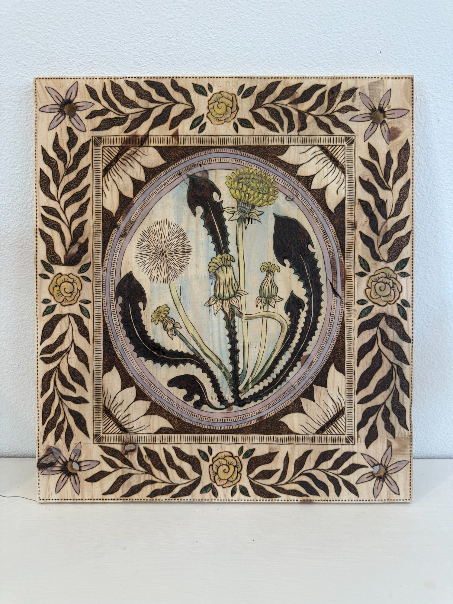Paige's handmade press named Luli — pyrography on hand-cut wood, 18 by 16 inches. Central dandelion medallion with seed head, bloom, and buds, surrounded by a heavily ornate burned border of roses, vines, and six-petal stars. Hand-colored in soft greens and yellows.