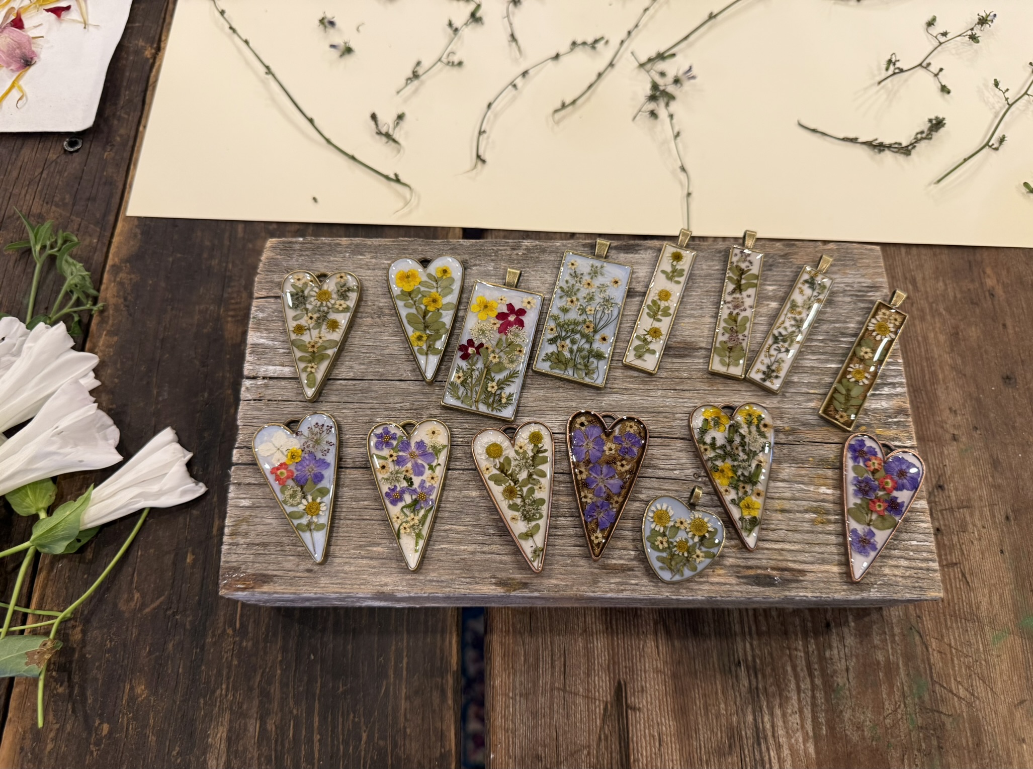 Collection of pressed flower pendants on barn wood — hearts and rectangles featuring purple verbena, yellow flowers, ferns and white blooms in clear and tinted resin bezels