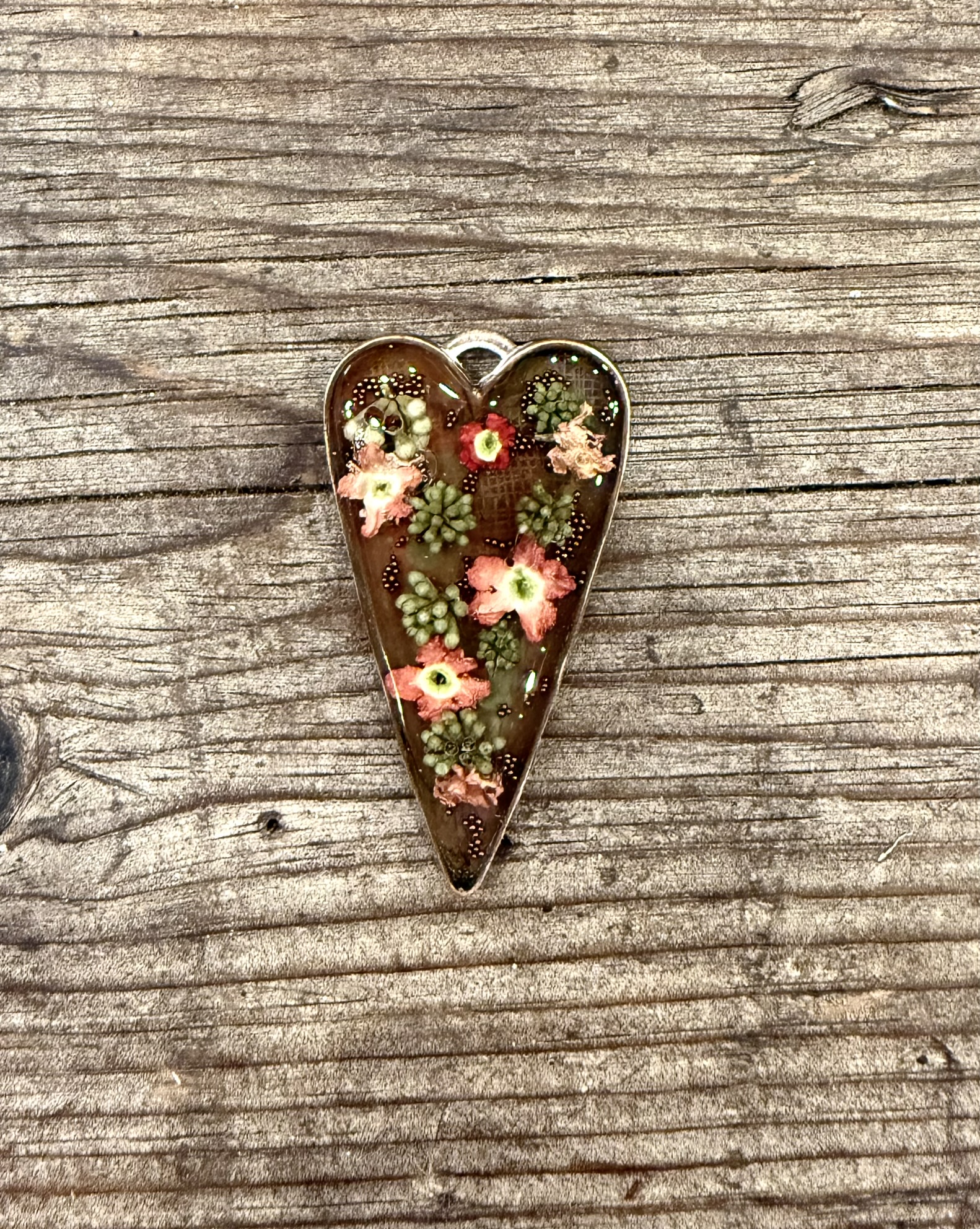 A dark resin heart pendant containing concentrated coral verbena flowers and mimosa tree blooms with green seed clusters — the flowers denser and deeper in color than they were living, set against the dark resin