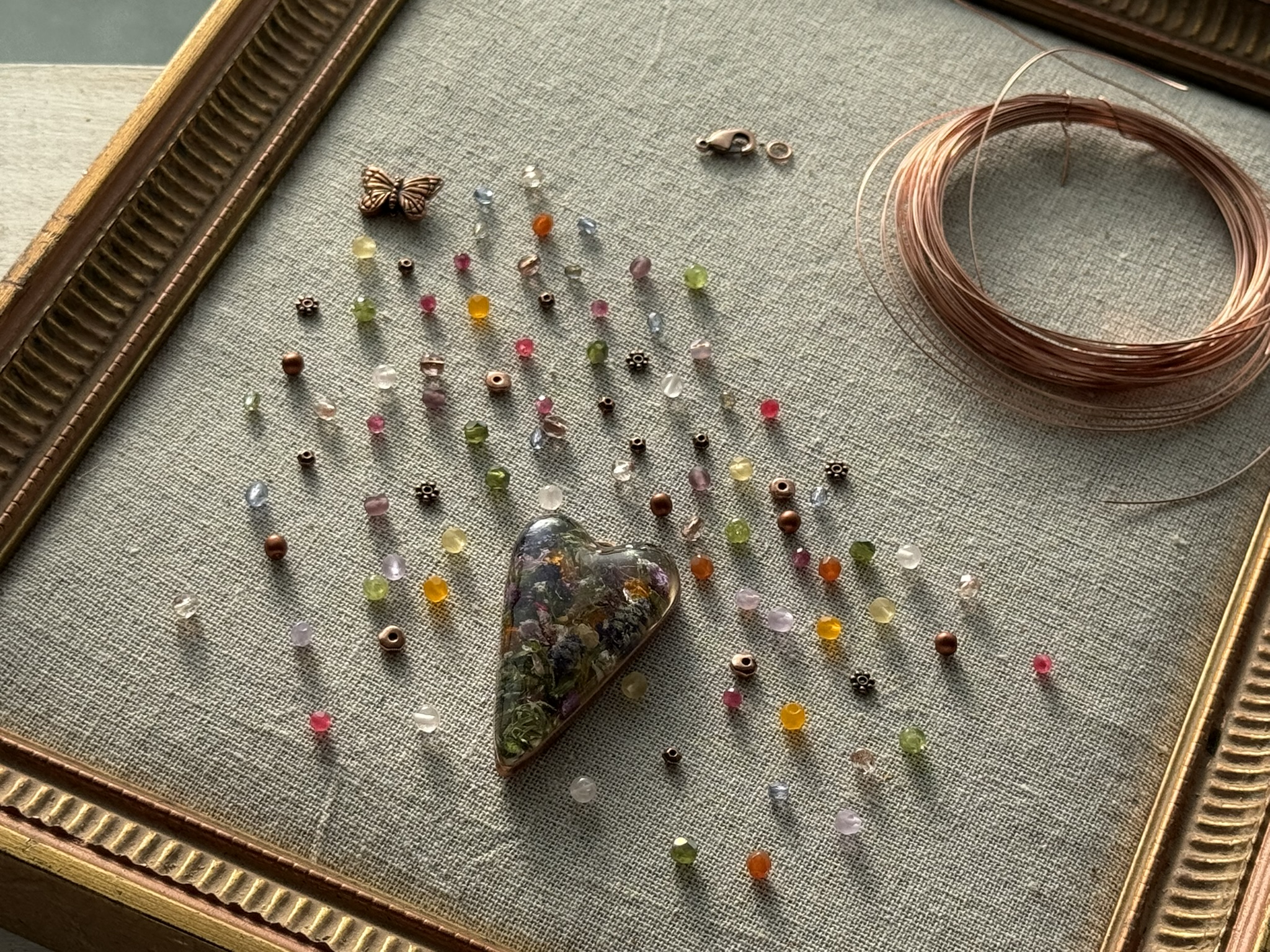 A heart pendant surrounded by every gemstone bead that will become its necklace — peridot, tourmaline, rose quartz, copper spacers, butterfly charm, wire coiled beside it — all laid out in a gold frame before assembly