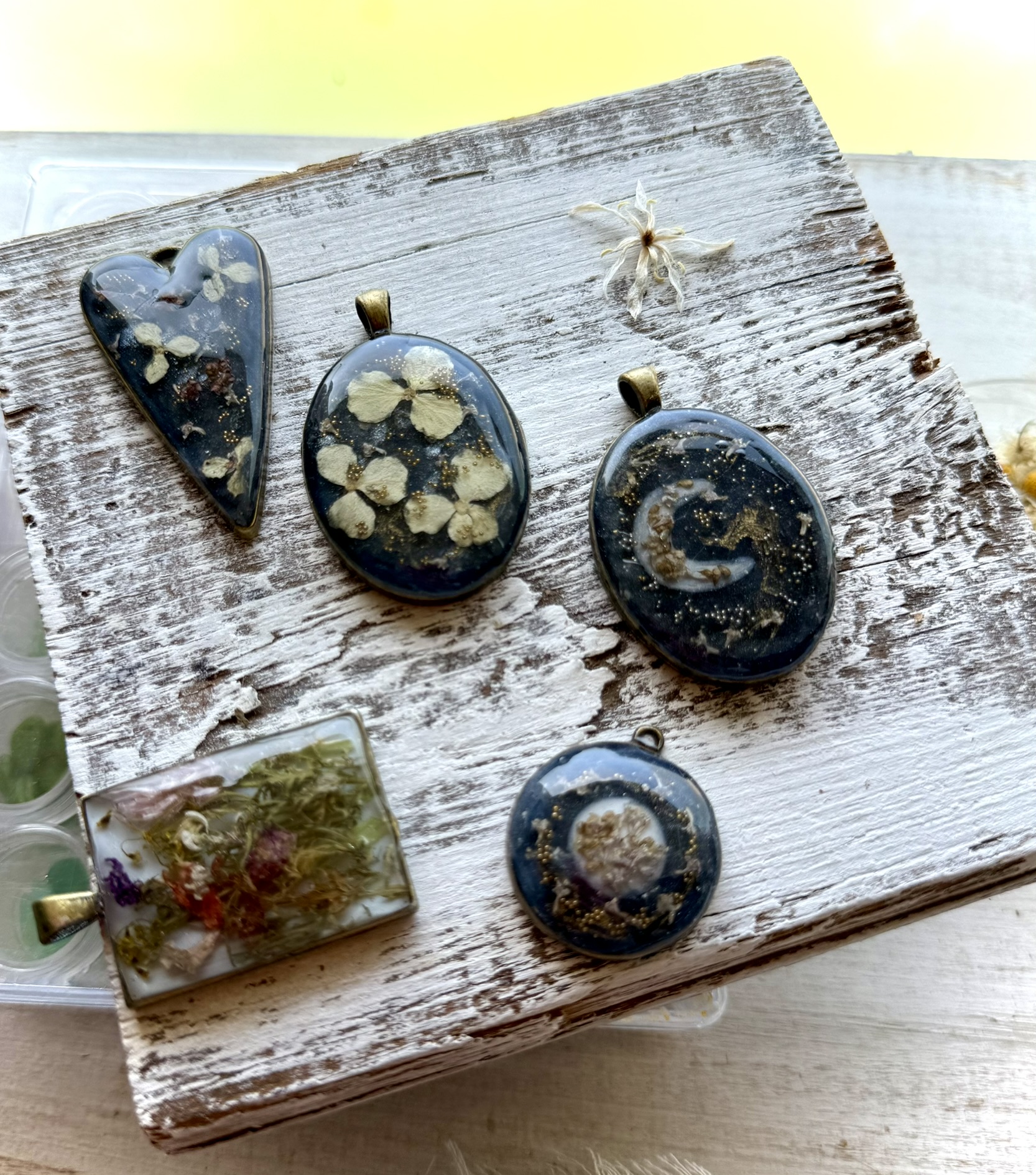 Five dark resin pendants on a whitewashed board — hearts and ovals with pressed flowers and gold glitter in deep resin, bronze bezels, a dried flower tucked at the corner