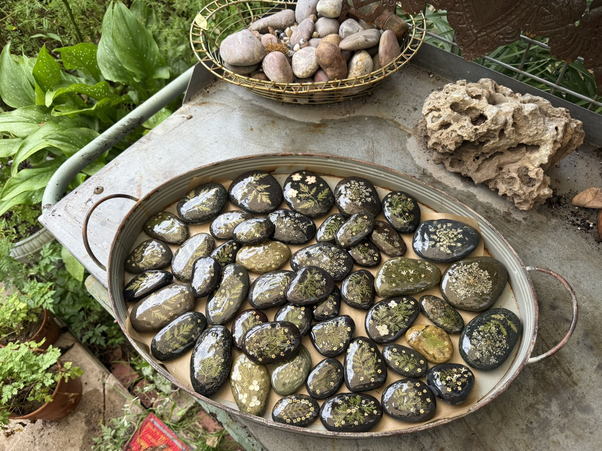 A vintage oval roasting pan packed with dark river stones each coated in resin over pressed botanicals — ferns and Queen Anne's lace gone golden-dark under resin, a basket of plain stones beside it