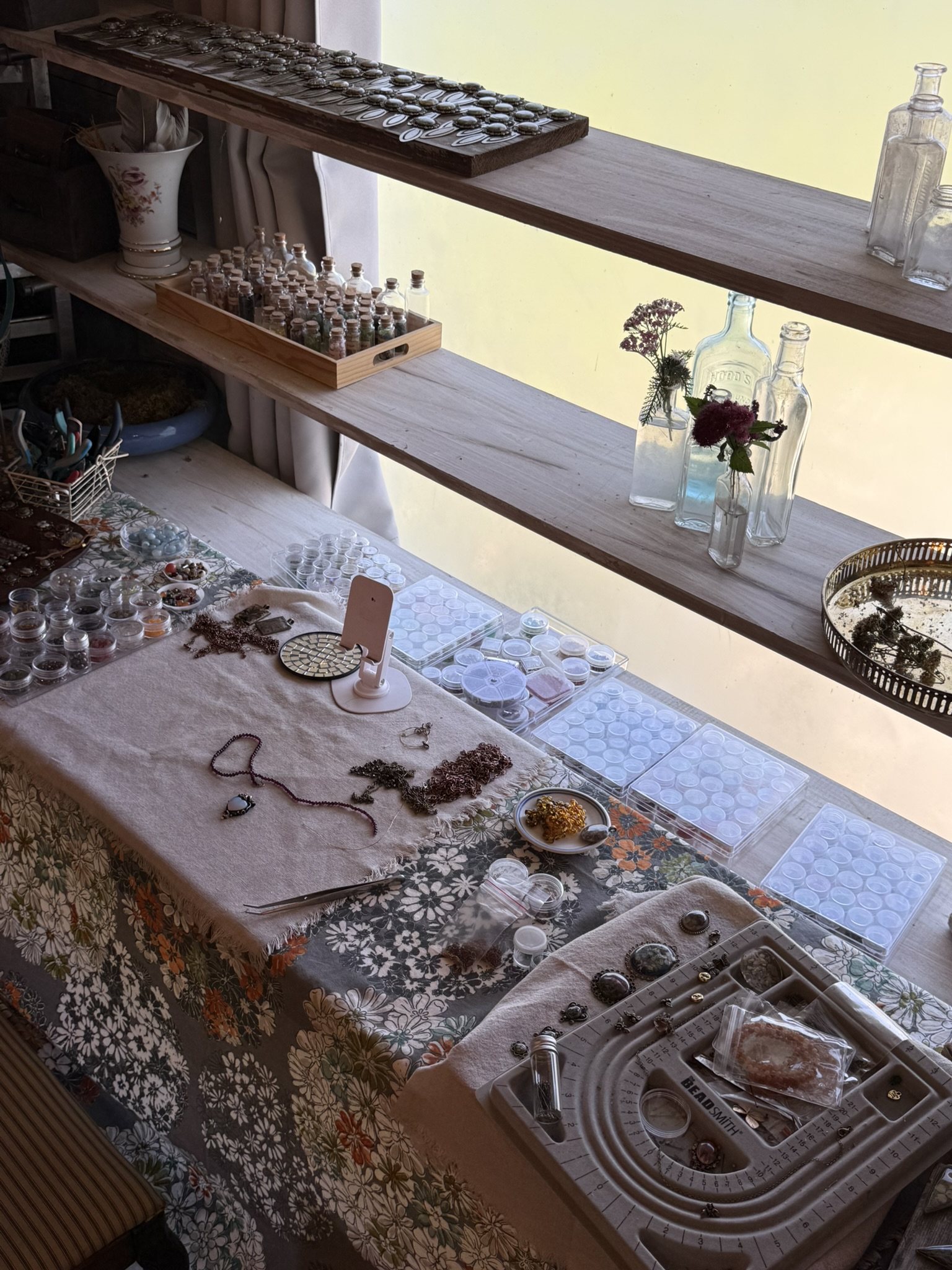 Studio bench from above — floral tablecloth covered in resin molds, beads, tweezers, necklaces in progress; shelves above lined with corked vials and vintage glass bottles; bezels on dark wood at the top