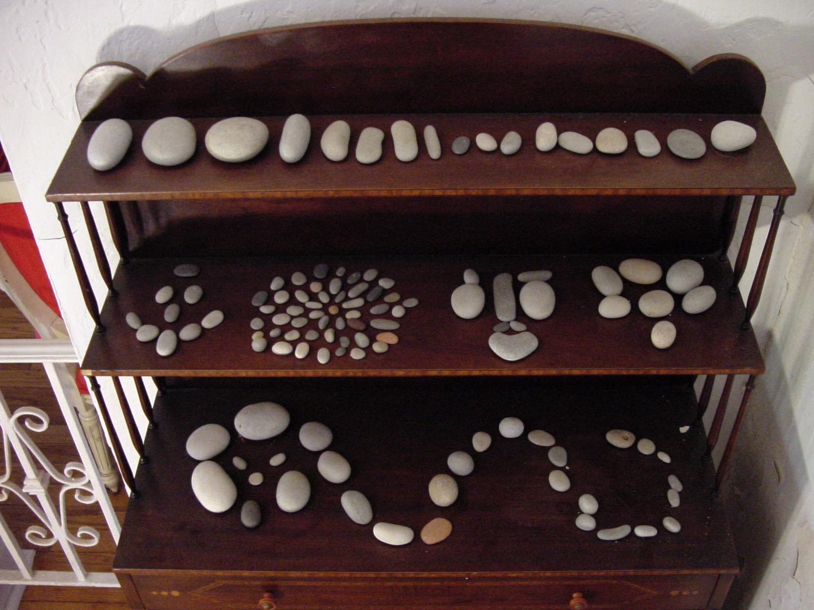 Close view of the three shelves: stones lined by size across the top, a mandala and a face of stones in the middle, flowing curves along the bottom
