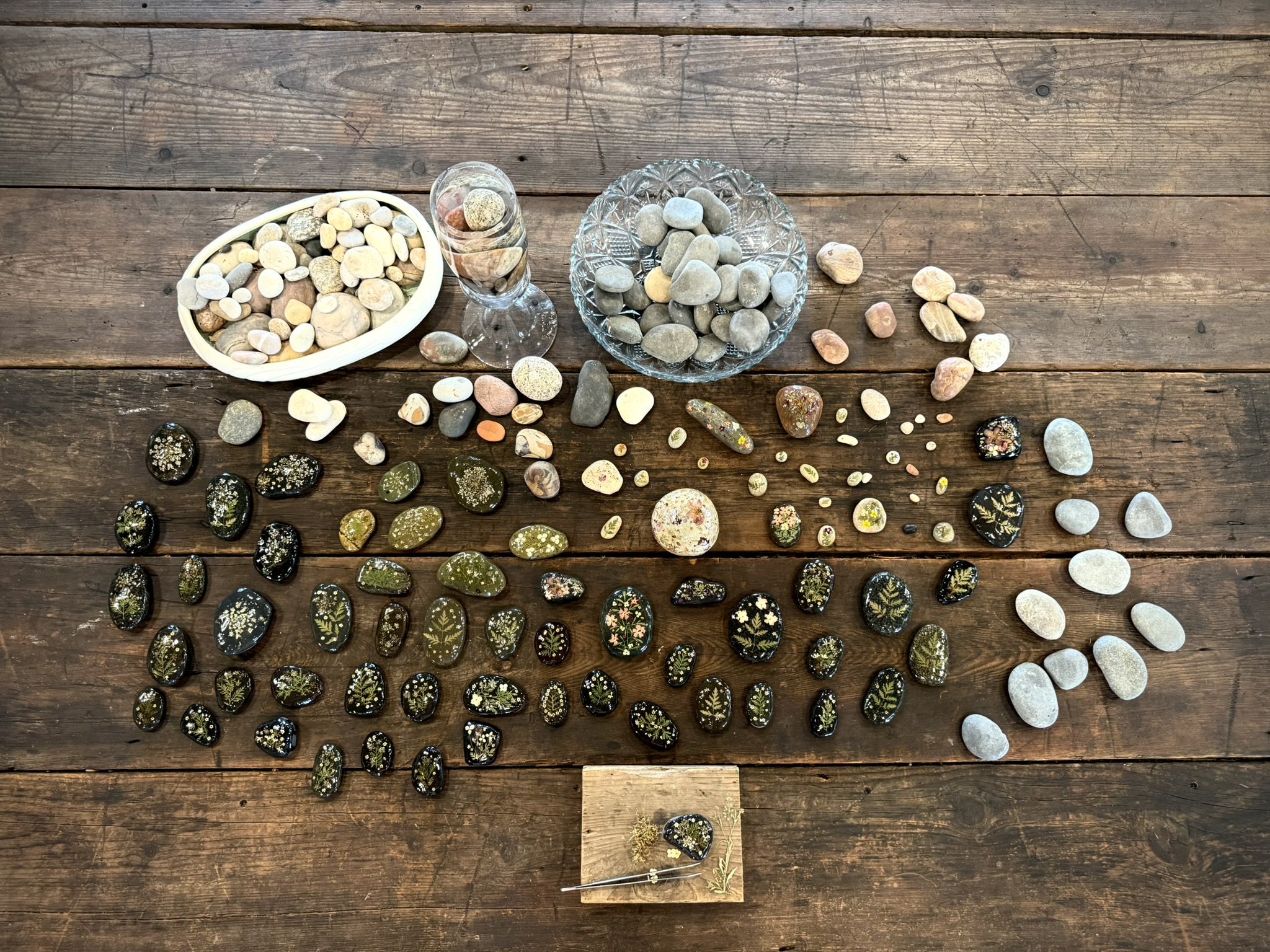 The full current collection: her natural river stones in white bowls and crystal dishes alongside the botanical pieces spread across an old wooden table — dozens of dark and olive-toned stones with pressed botanicals sealed in resin