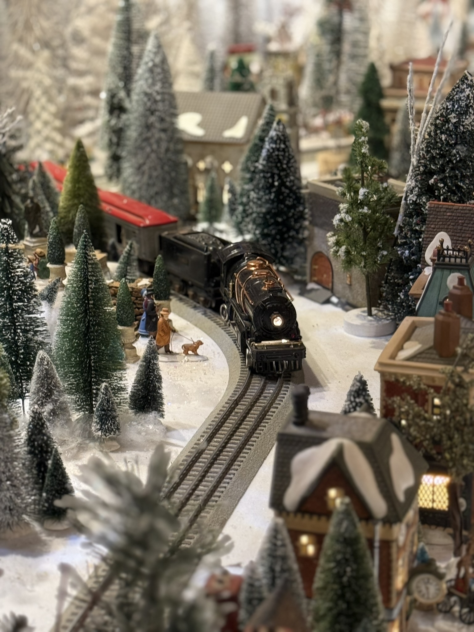 Christmas train village close-up: black steam locomotive curving through snow-covered buildings, miniature figures walking a dog beside the track