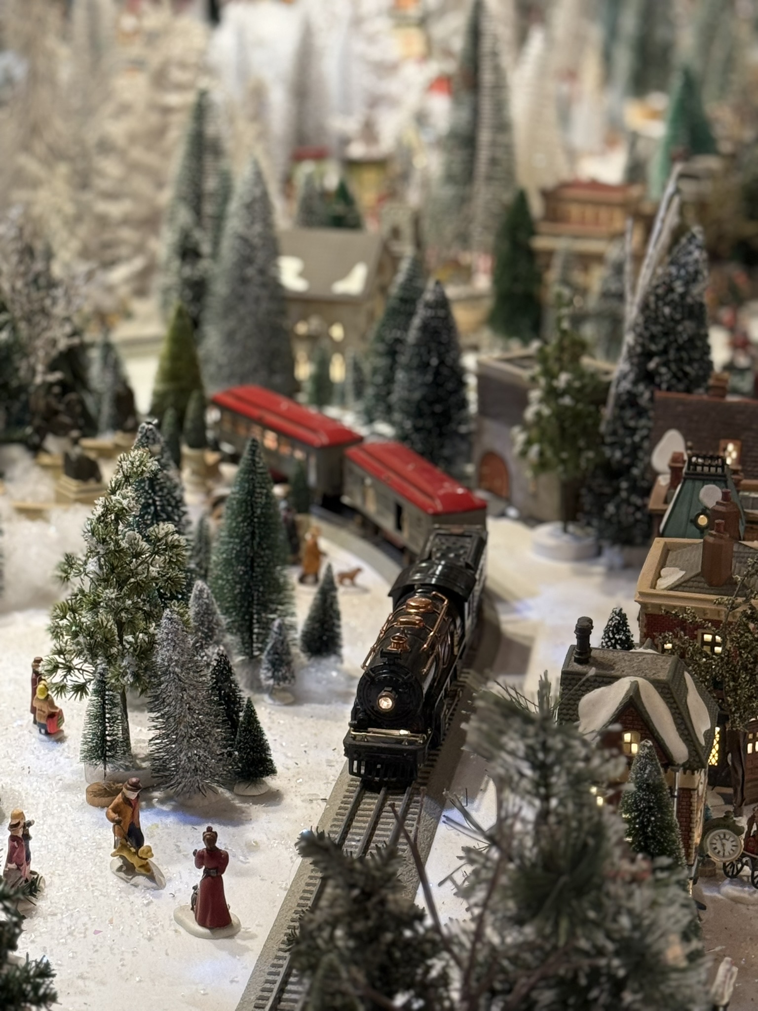 Christmas train village wider view: red passenger car rounding a bend, tiny figures and snow-covered storefronts lit from within
