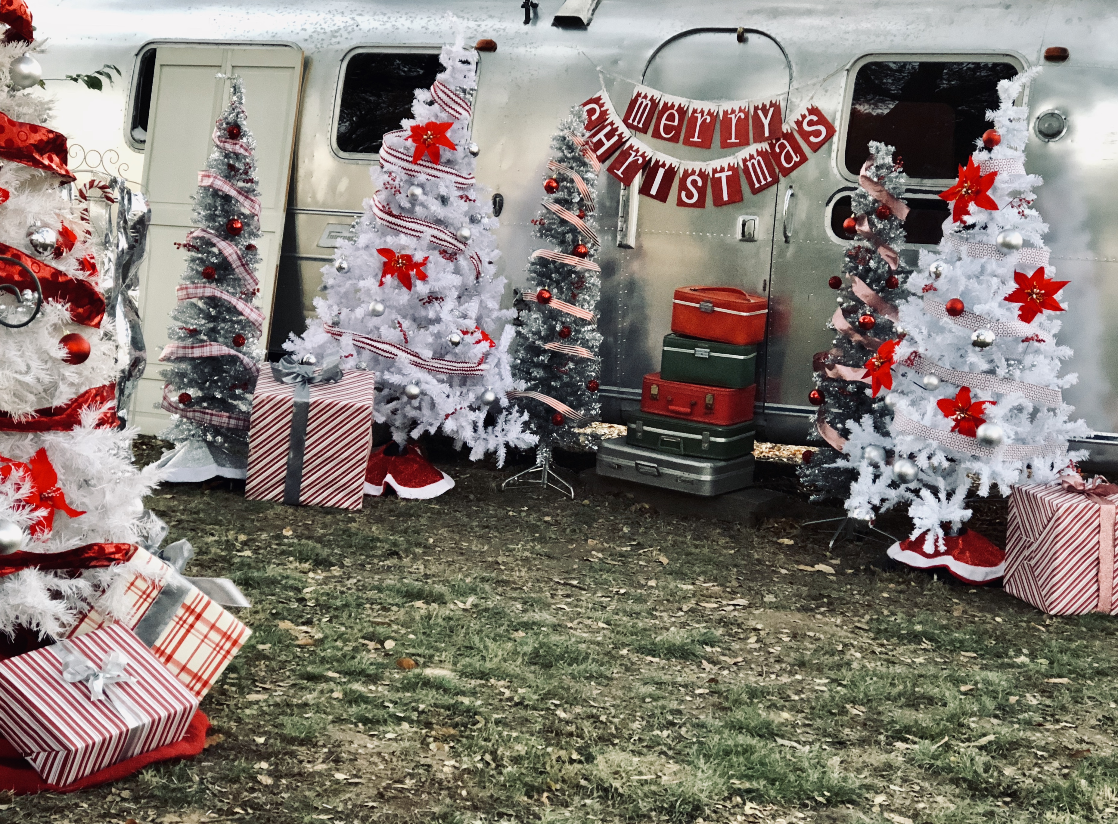 Vintage Airstream trailer as holiday photo backdrop, flanked by white Christmas trees with red ornaments and stacked vintage luggage, wrapped gifts on the lawn