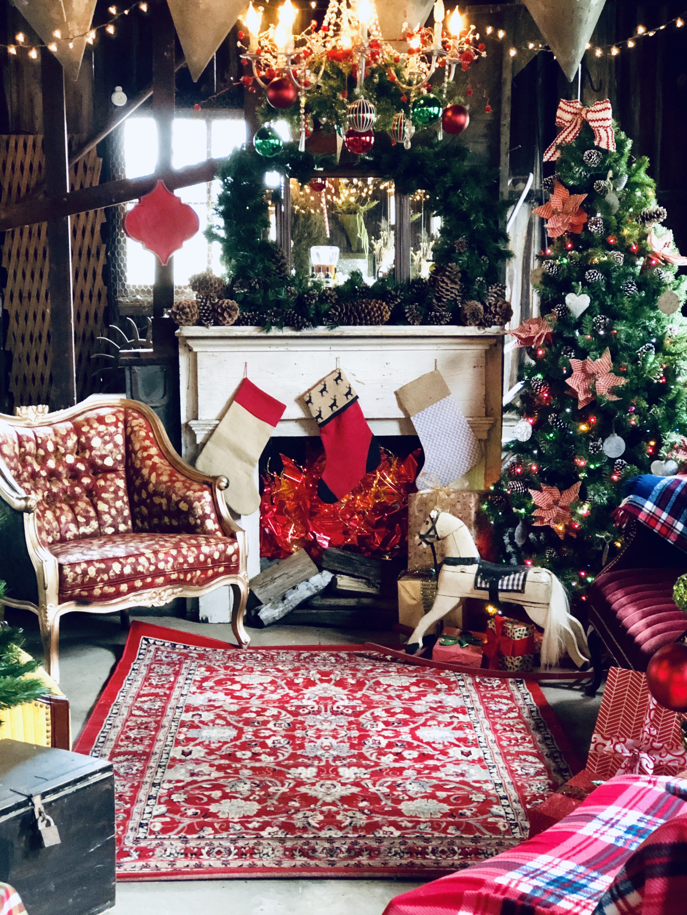 Barn fireplace vignette with stockings, a rocking horse, vintage armchair in red print fabric, chandelier draped in ornaments and berries