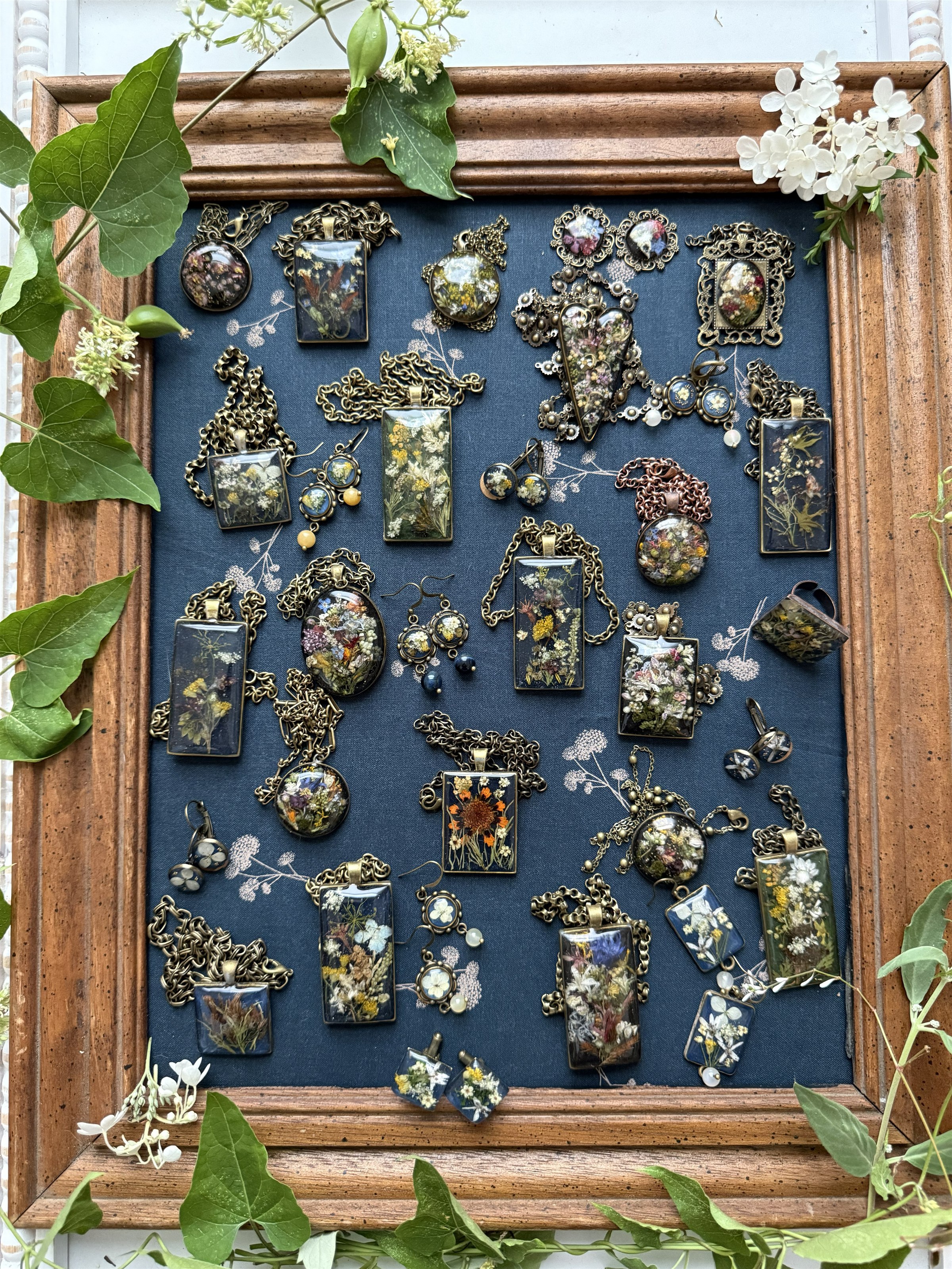 Floral Alchemy pieces arranged on dark botanical fabric in a wood frame, surrounded by living foliage