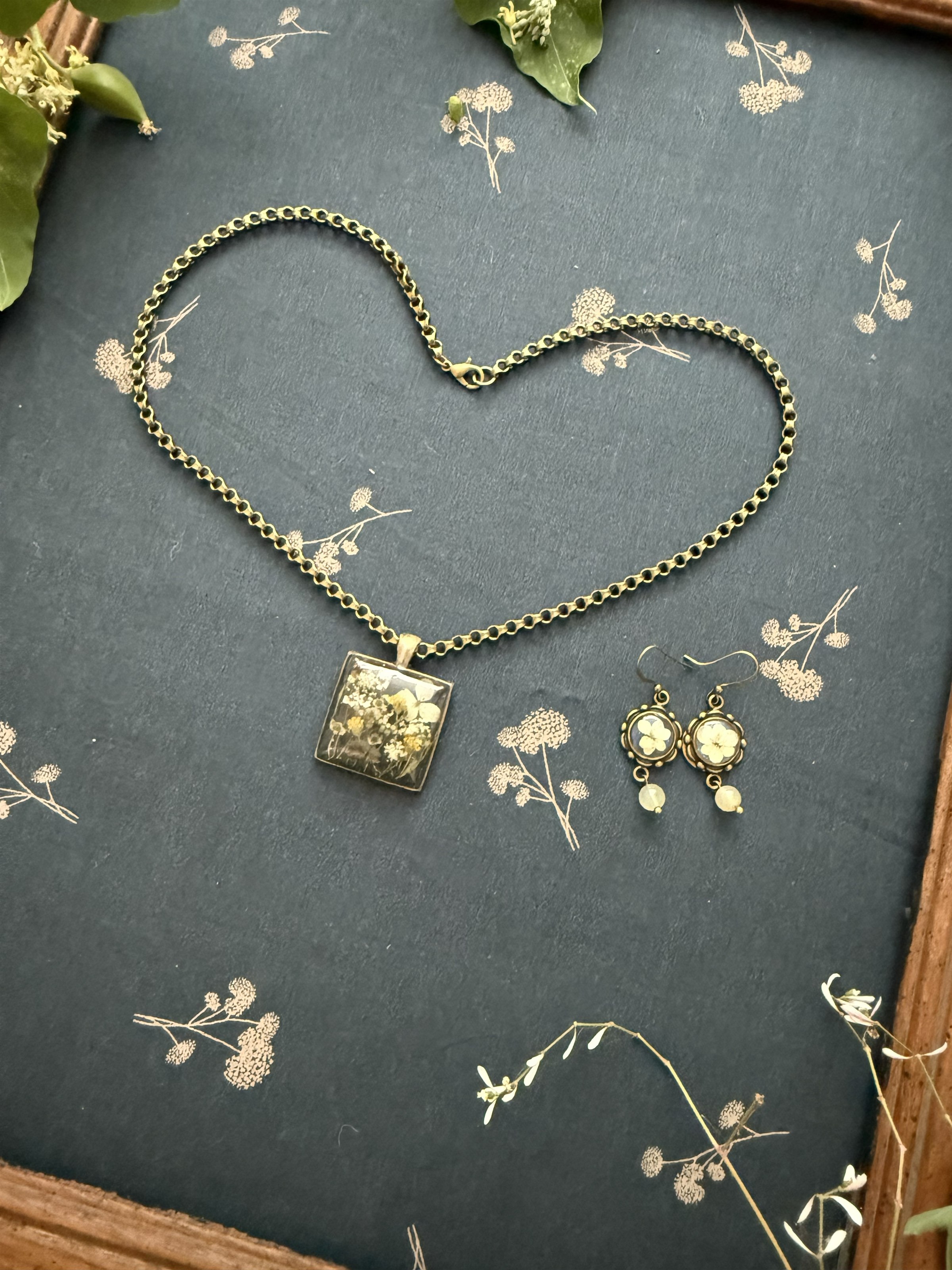 A square botanical pendant and earrings on a heart-shaped chain, intimate and tender on blue botanical fabric
