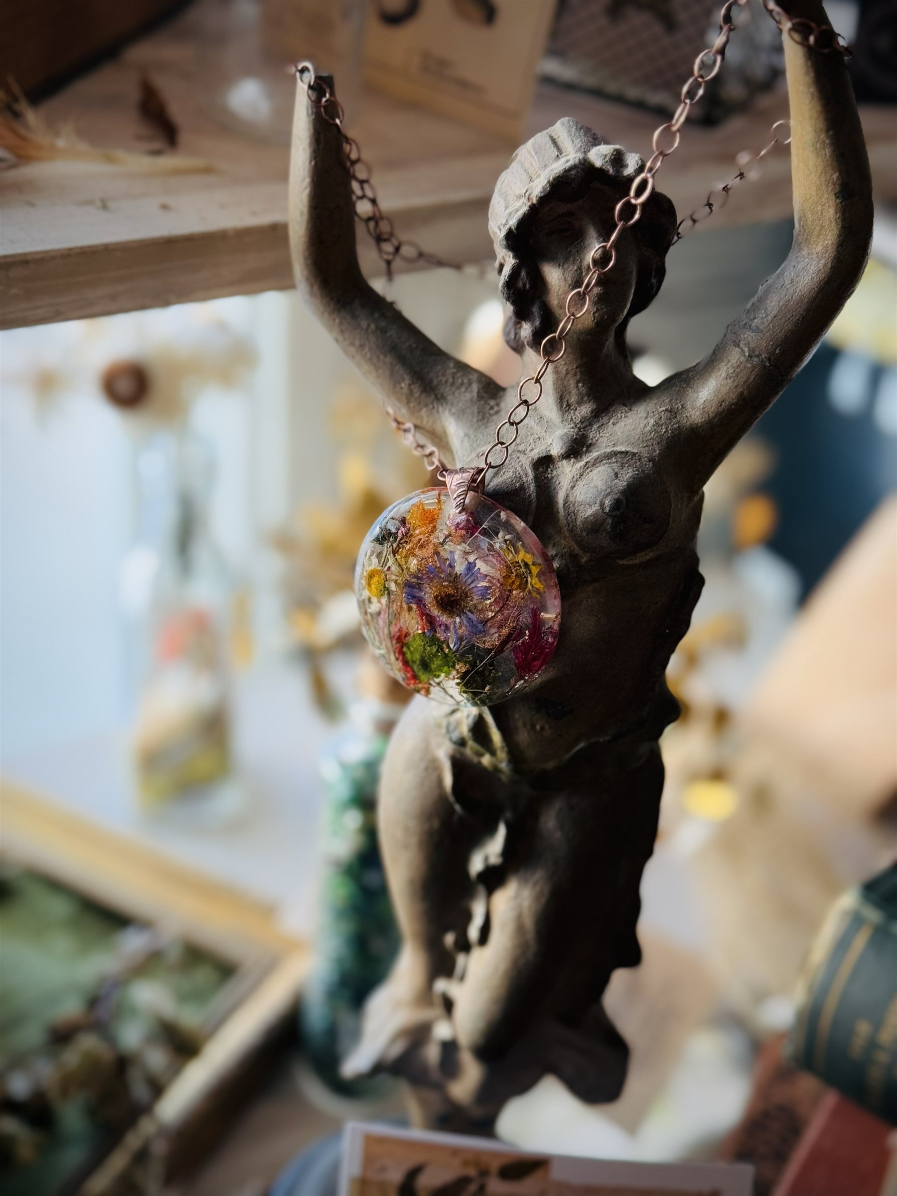 A botanical pendant held aloft by a bronze figurine with arms raised — Floral Alchemy