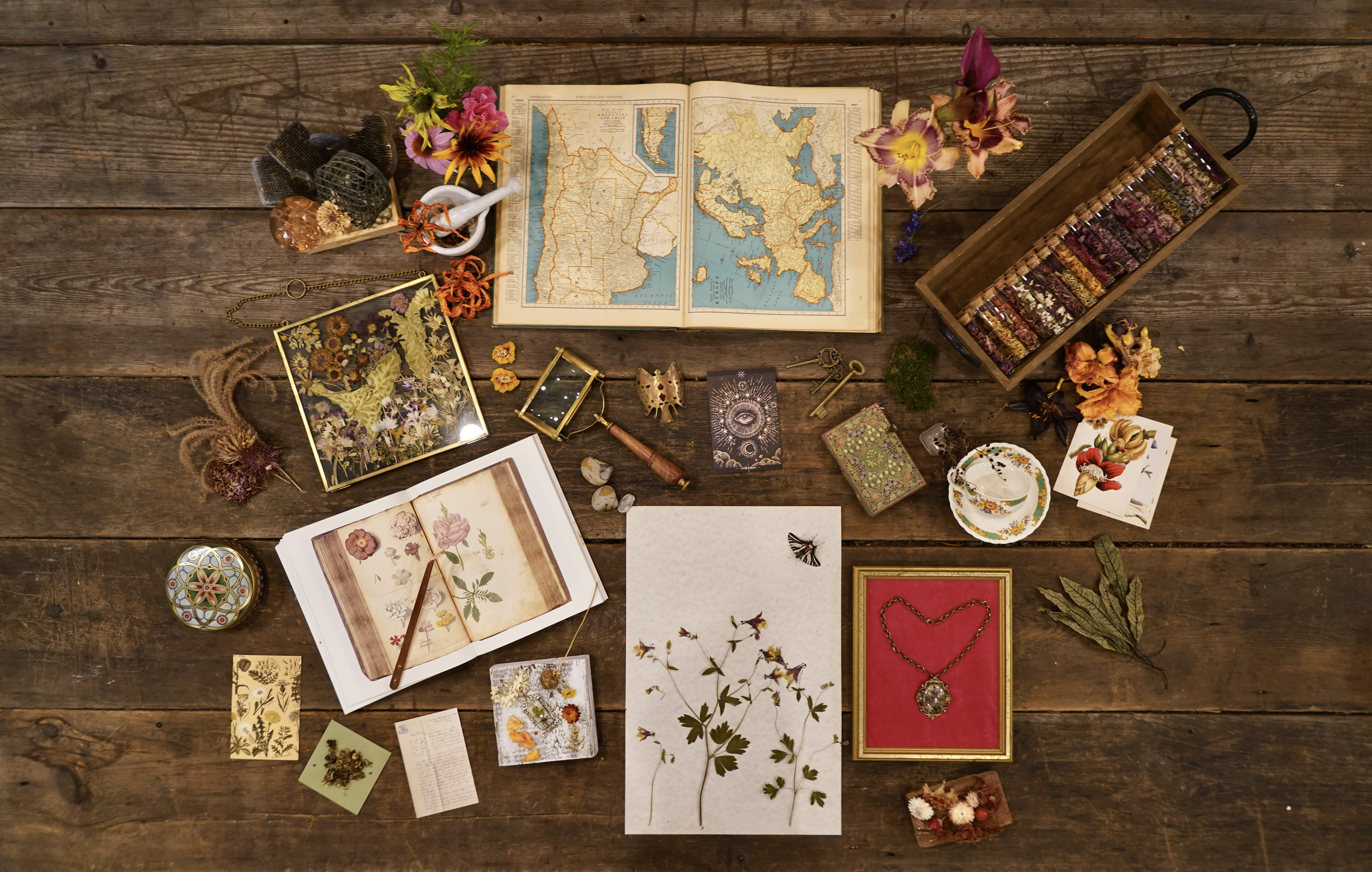 A table of curated objects — maps, keys, pressed flowers, botanical books, specimen vials, a finished piece — each one a door