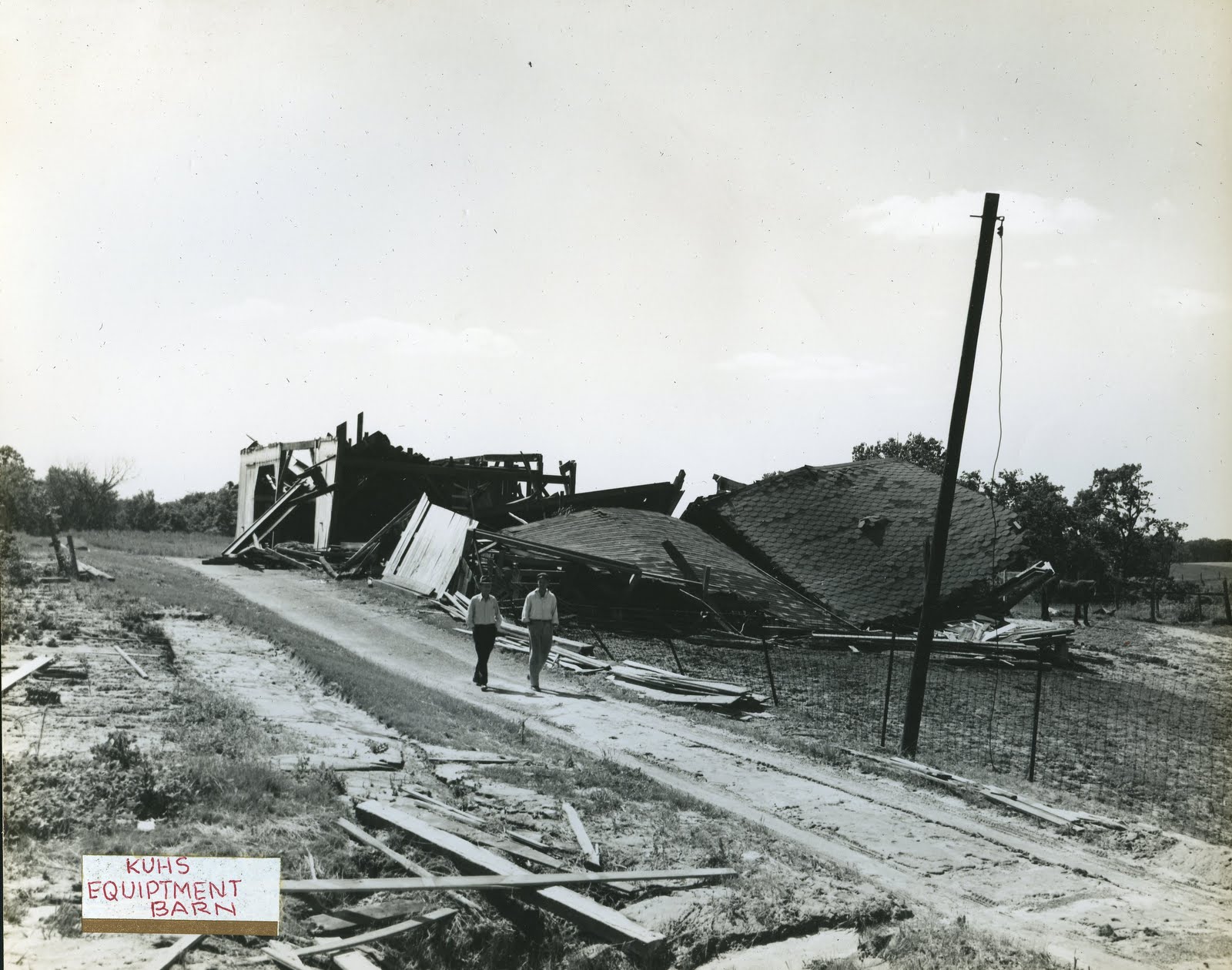 The equipment barn after the 1949 tornado — completely destroyed, flattened, two men walking past the rubble